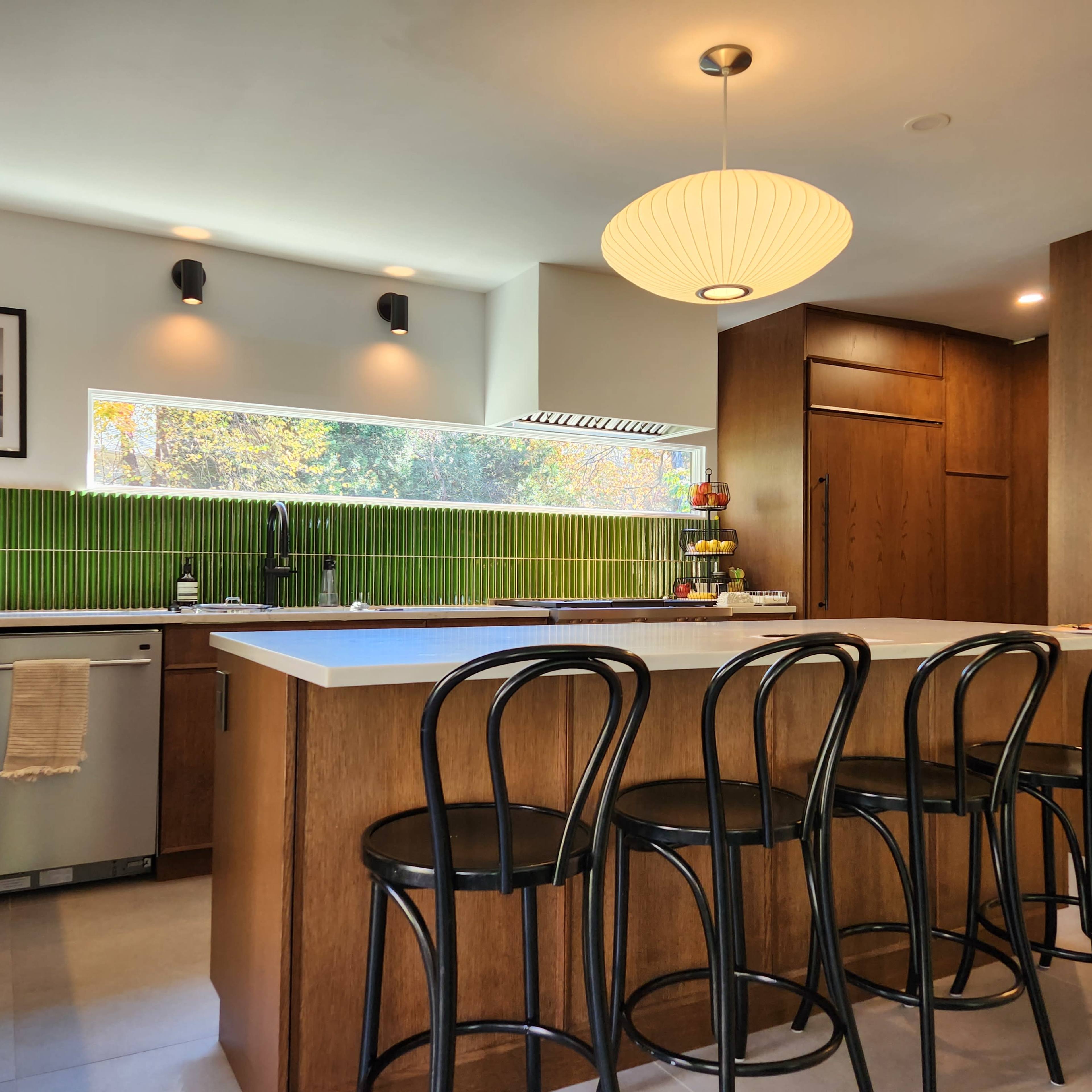 A modern kitchen features a large island with bar stools, wooden cabinetry, and a green tile backsplash.