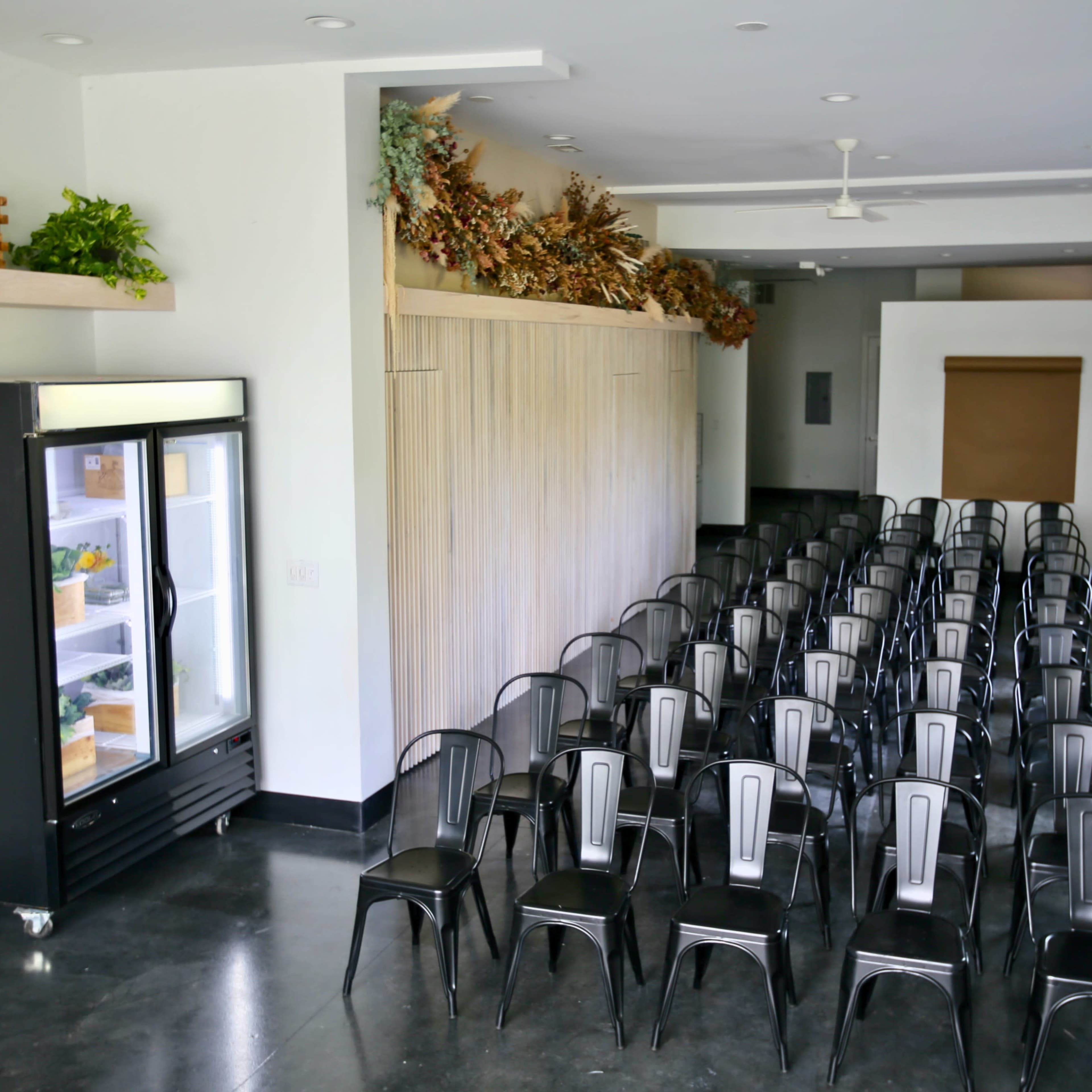 A spacious room features a row of black chairs facing a wall adorned with framed photographs, alongside a glass-front refrigerator and decorative elements on the shelves.