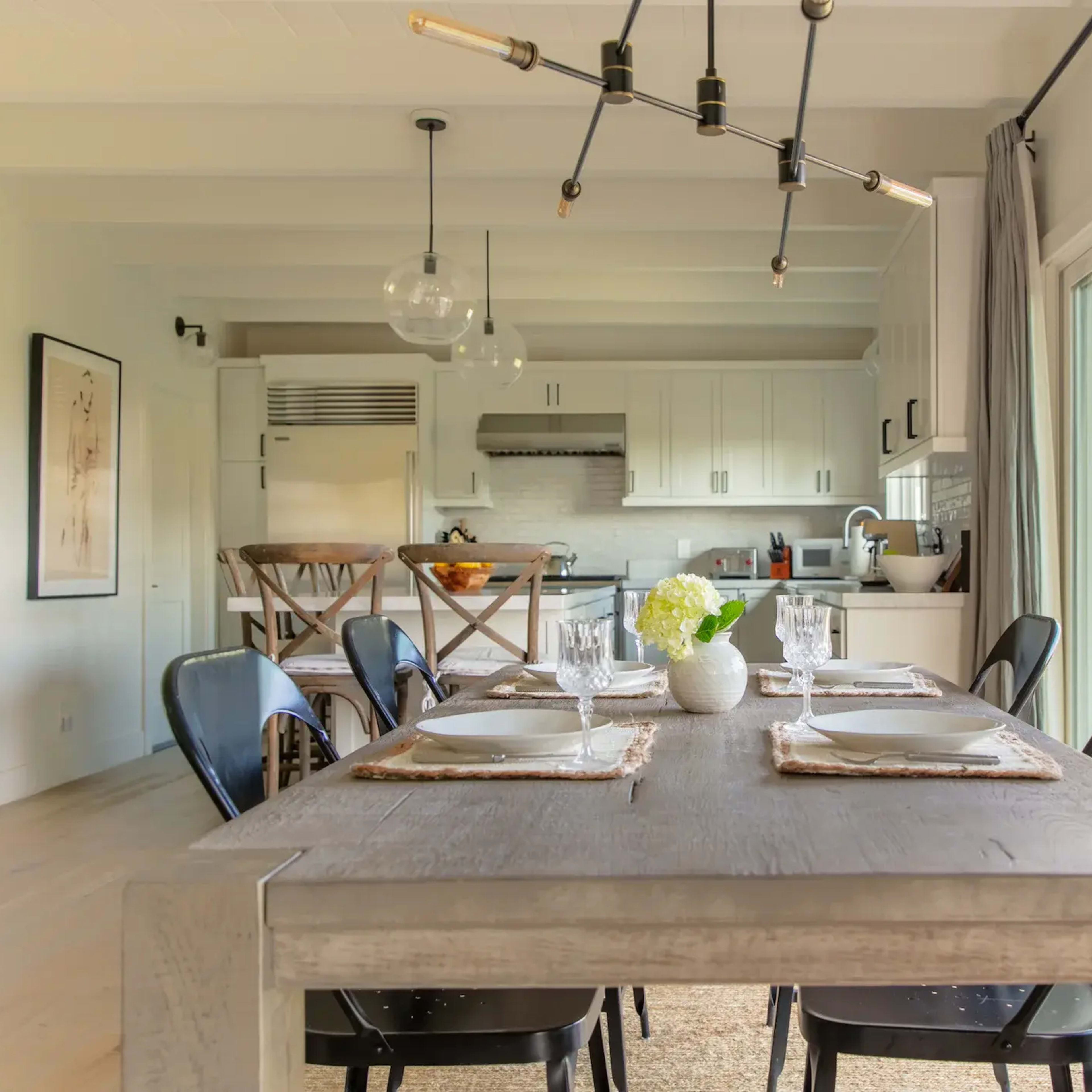 A wooden dining table with seating for six is set in a bright room that connects to a modern kitchen through large windows.