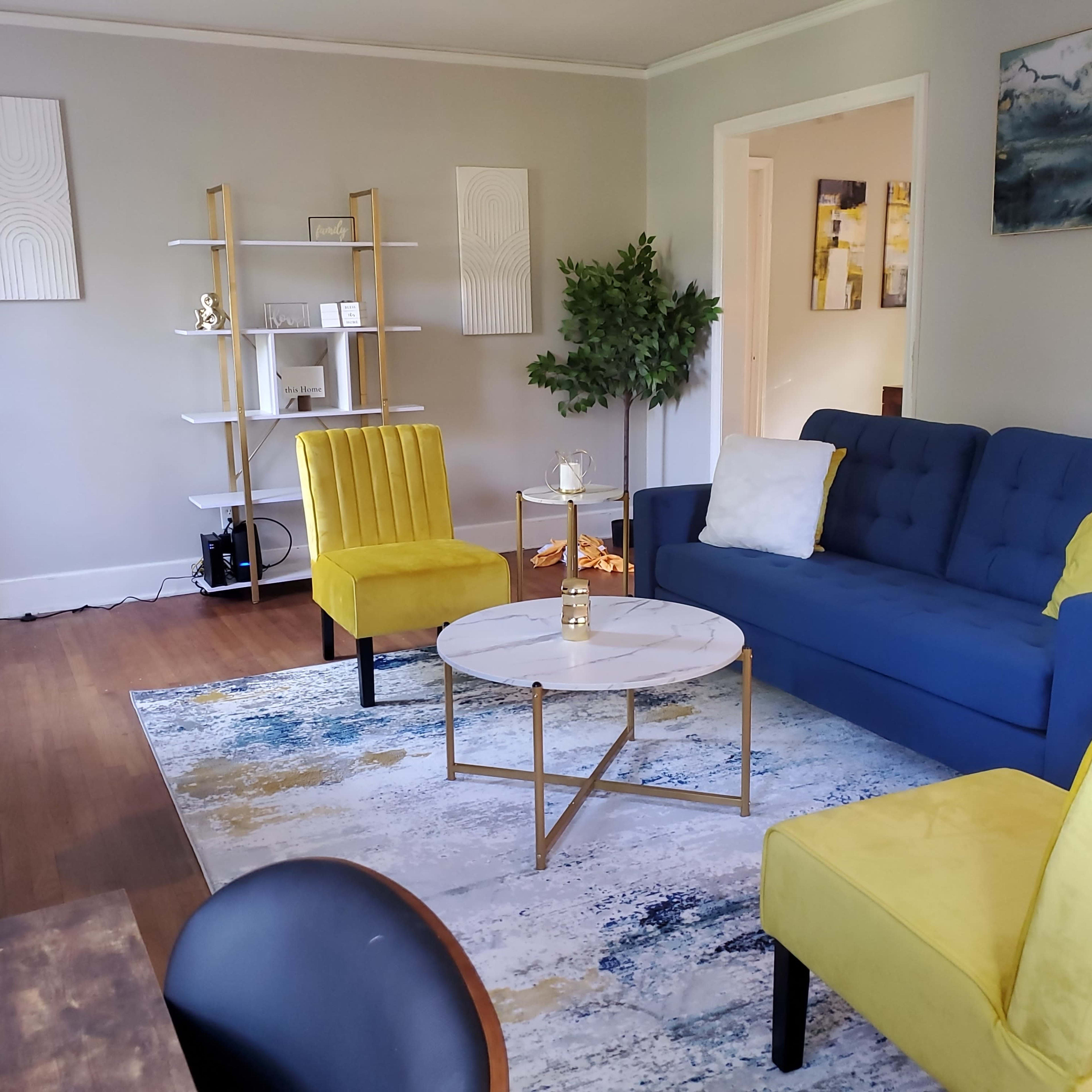 The living room features a blue sofa, two yellow chairs, a round coffee table, and a decorative rug, all arranged around a bookshelf and a potted plant.
