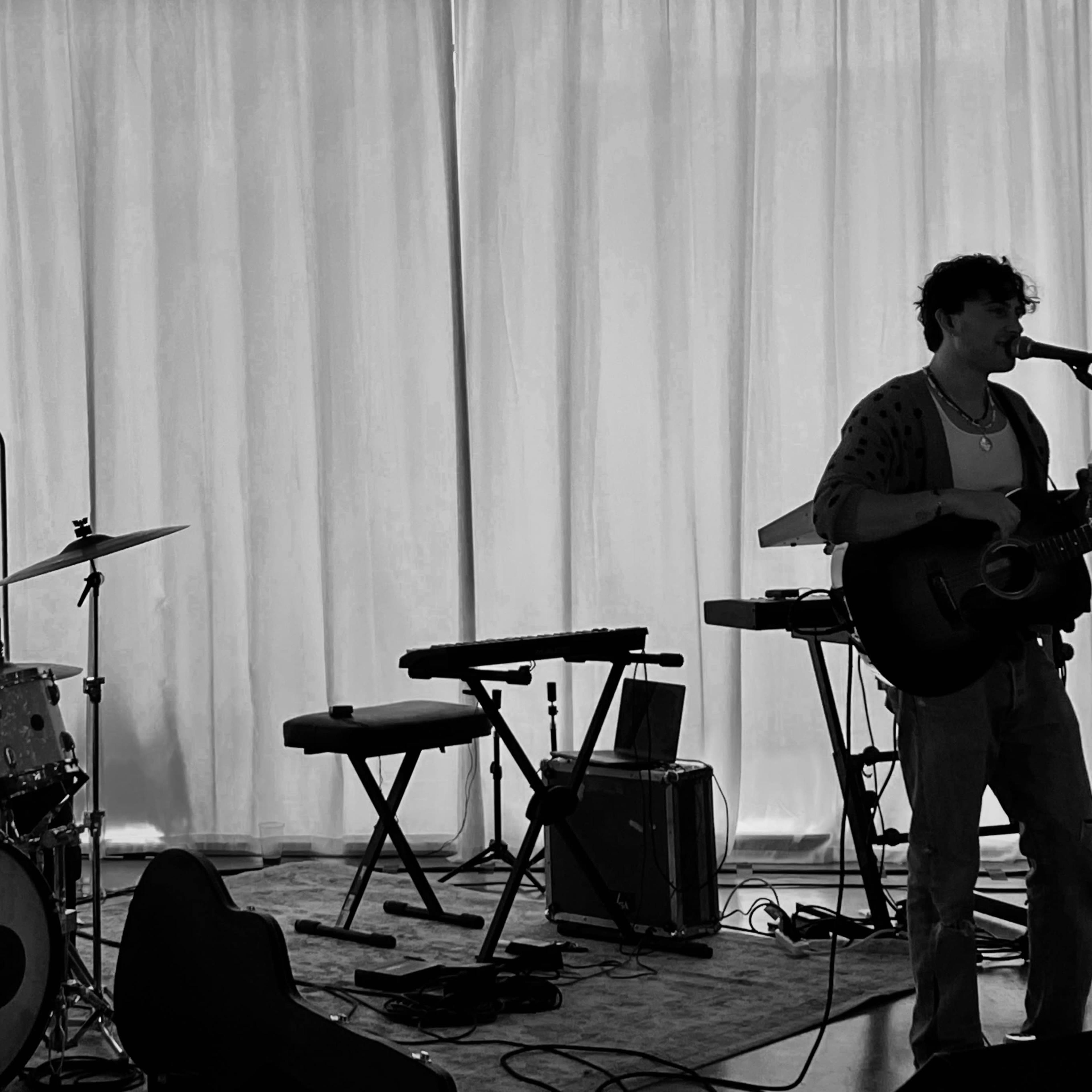 A musician performs with a guitar in front of a drum set and keyboard on a stage.