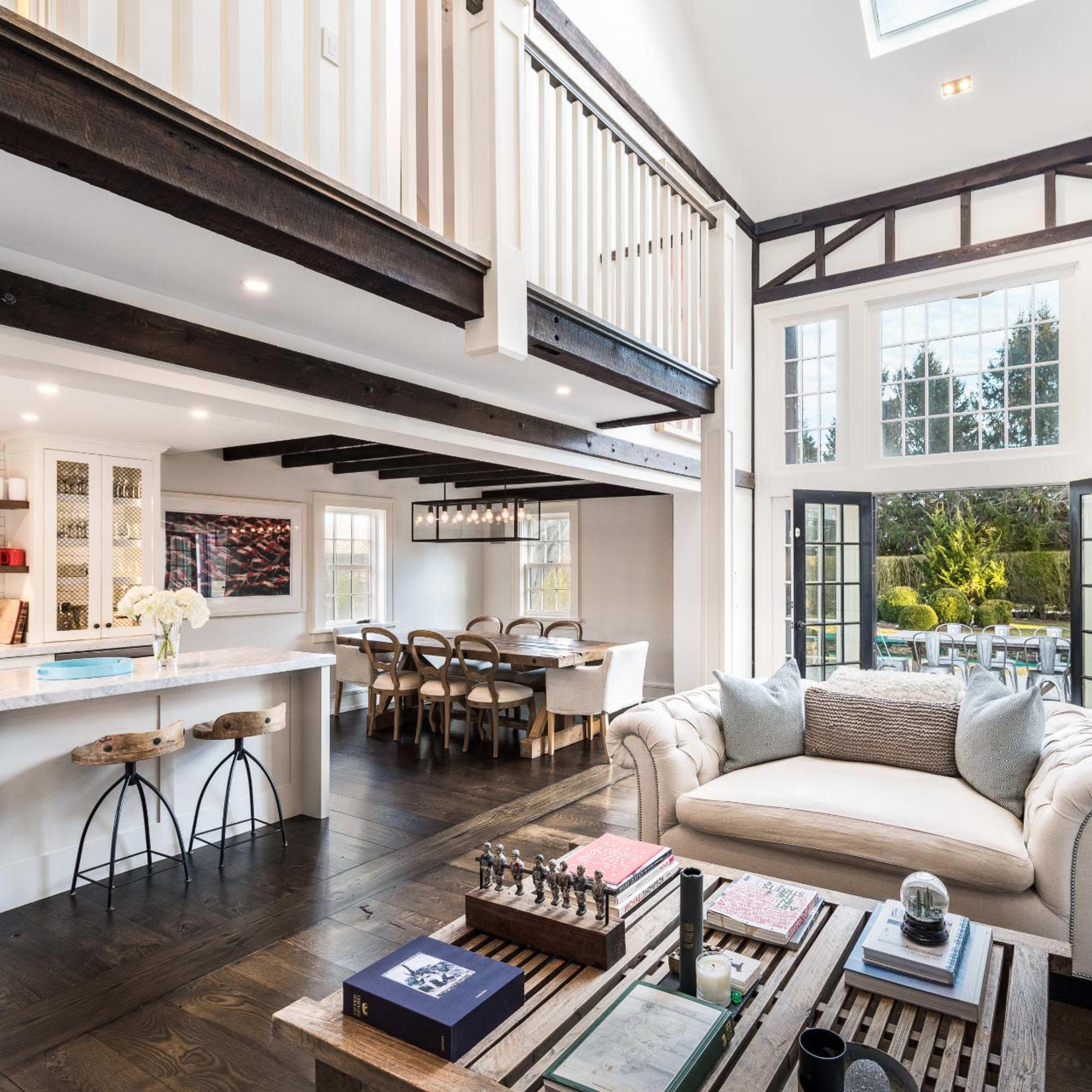 A spacious open-plan living area features a cream-colored couch, a coffee table with chess pieces, and large windows that open to a garden, complemented by a dining area in the background.