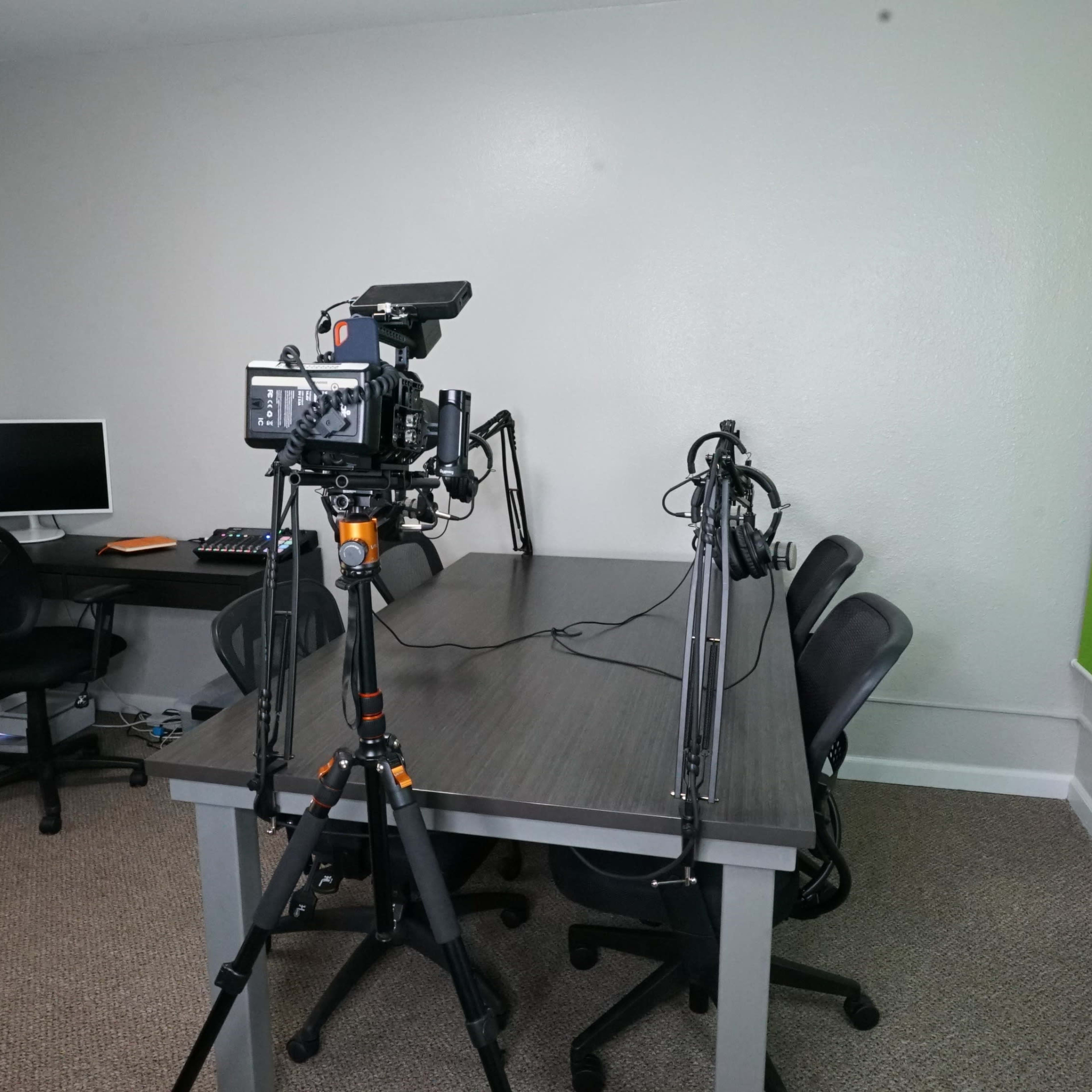 A camera is set up on a tripod facing a table with three chairs in a room featuring a green wall, a computer, and a window with blinds.
