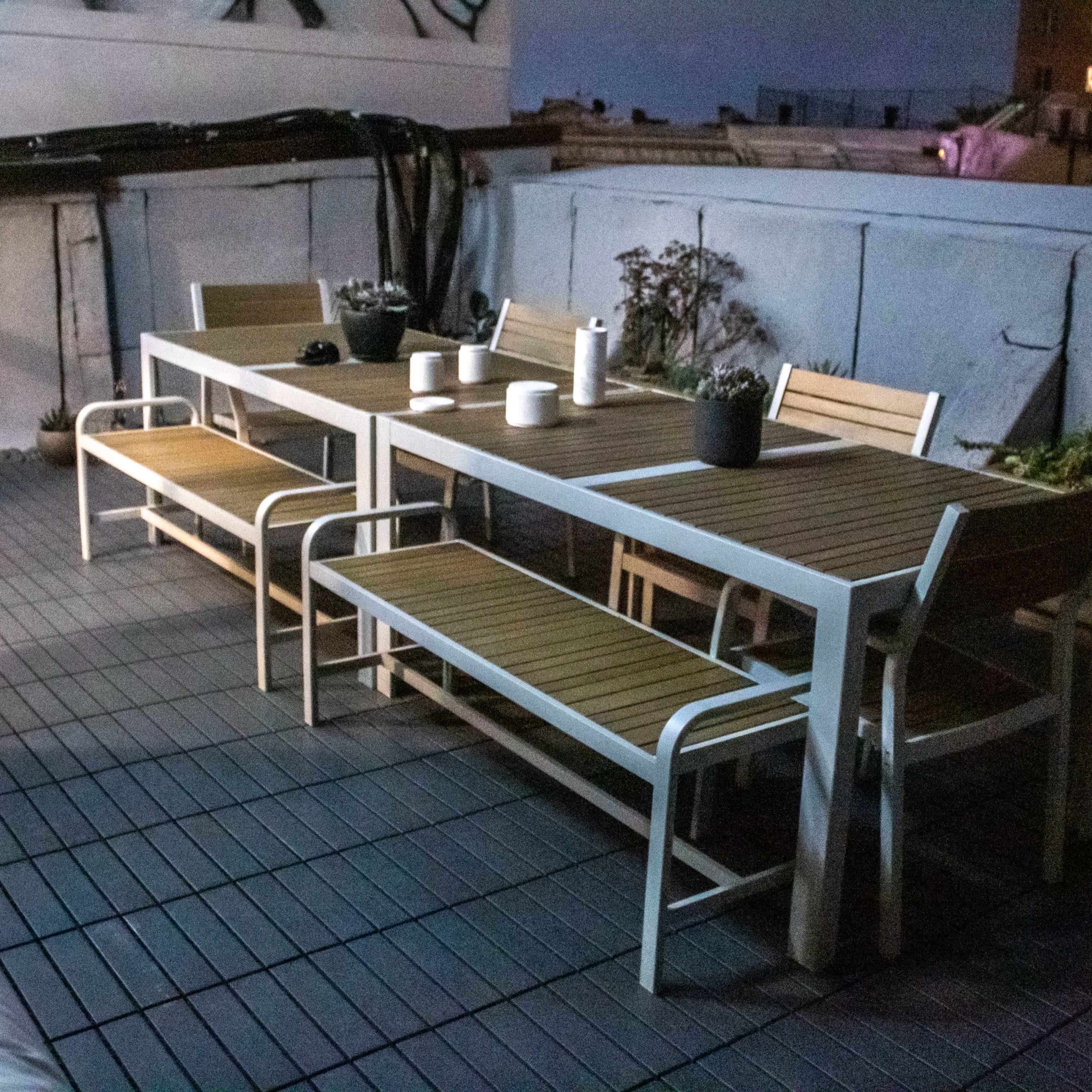 A wooden dining table with benches is set on a rooftop terrace, surrounded by potted plants and illuminated by ambient night lighting.