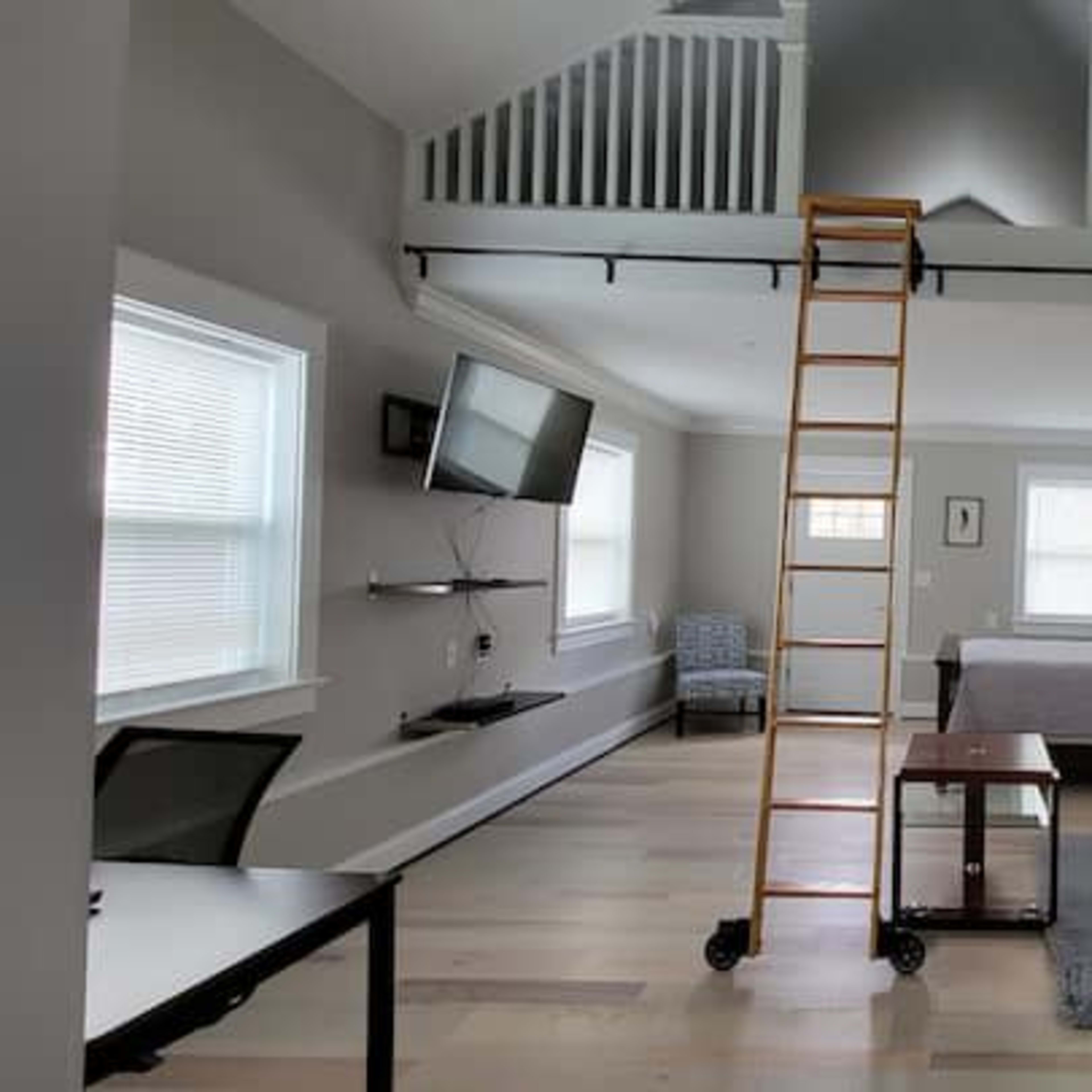 A modern room with a lofted area, featuring a bed, a yellow sofa, a desk, and a ladder leading to the upper level.