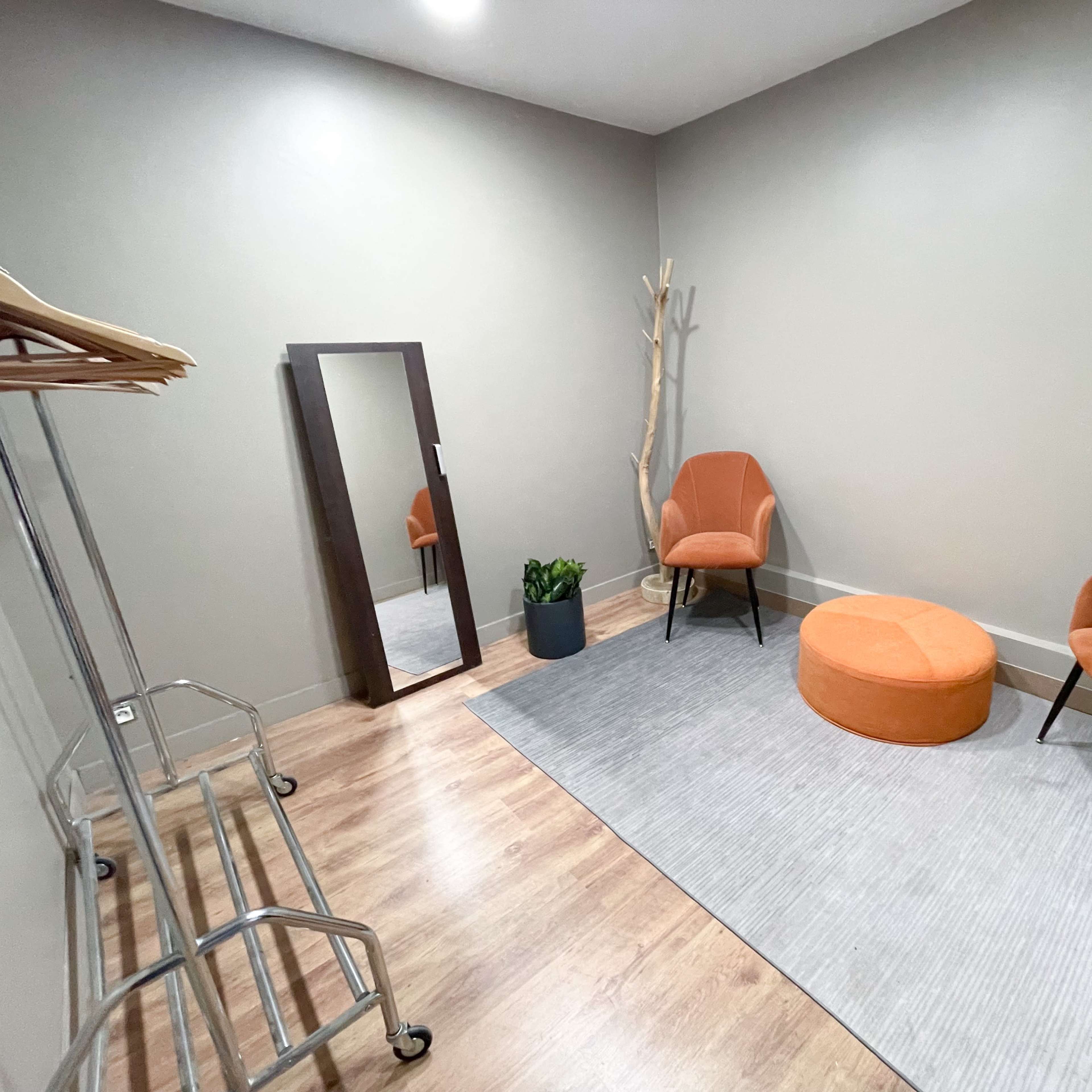 A small room with two orange chairs, a round orange ottoman, a full-length mirror, a plant, and a clothing rack against a gray wall.