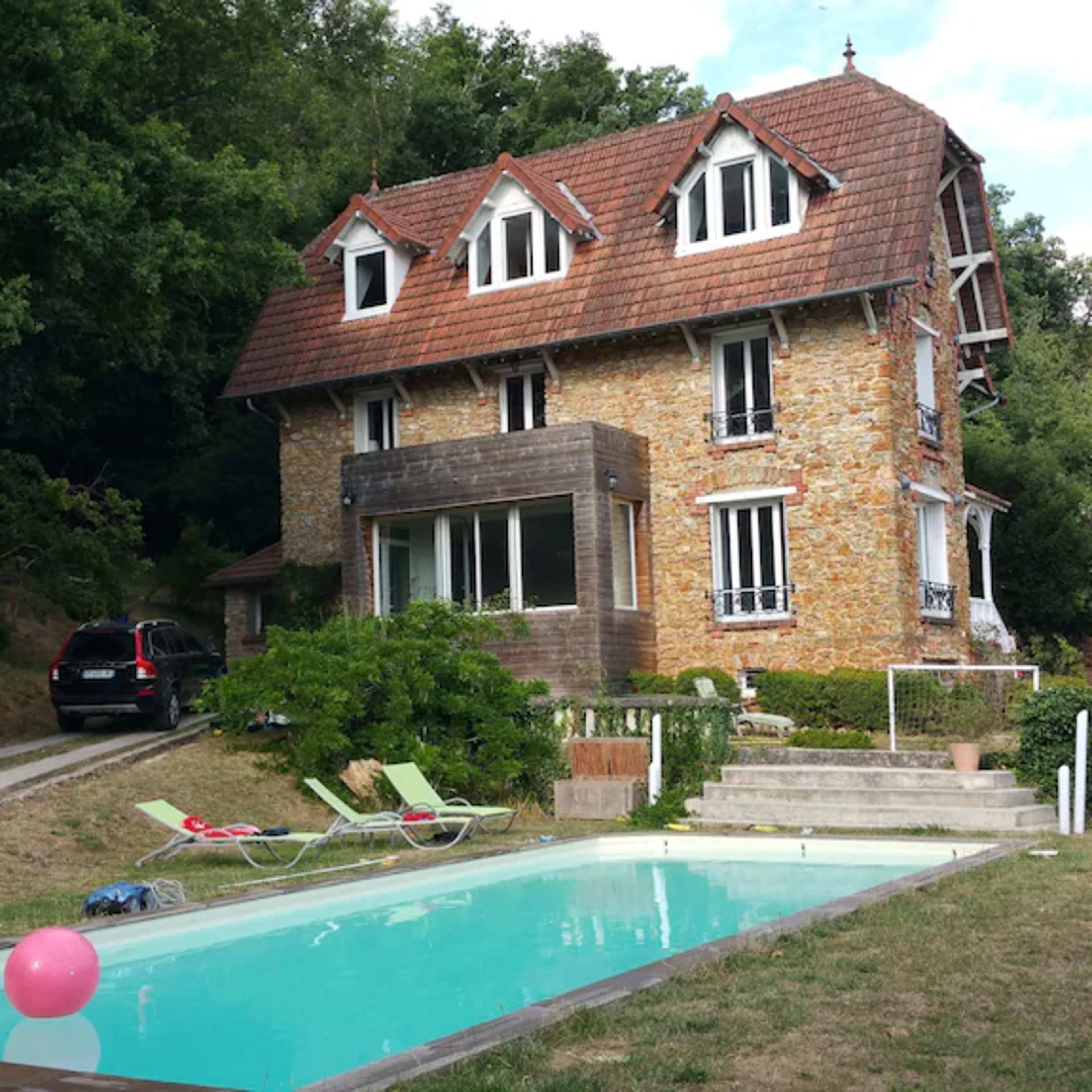 A three-story stone house with a sloped roof is situated near a swimming pool and surrounded by greenery.