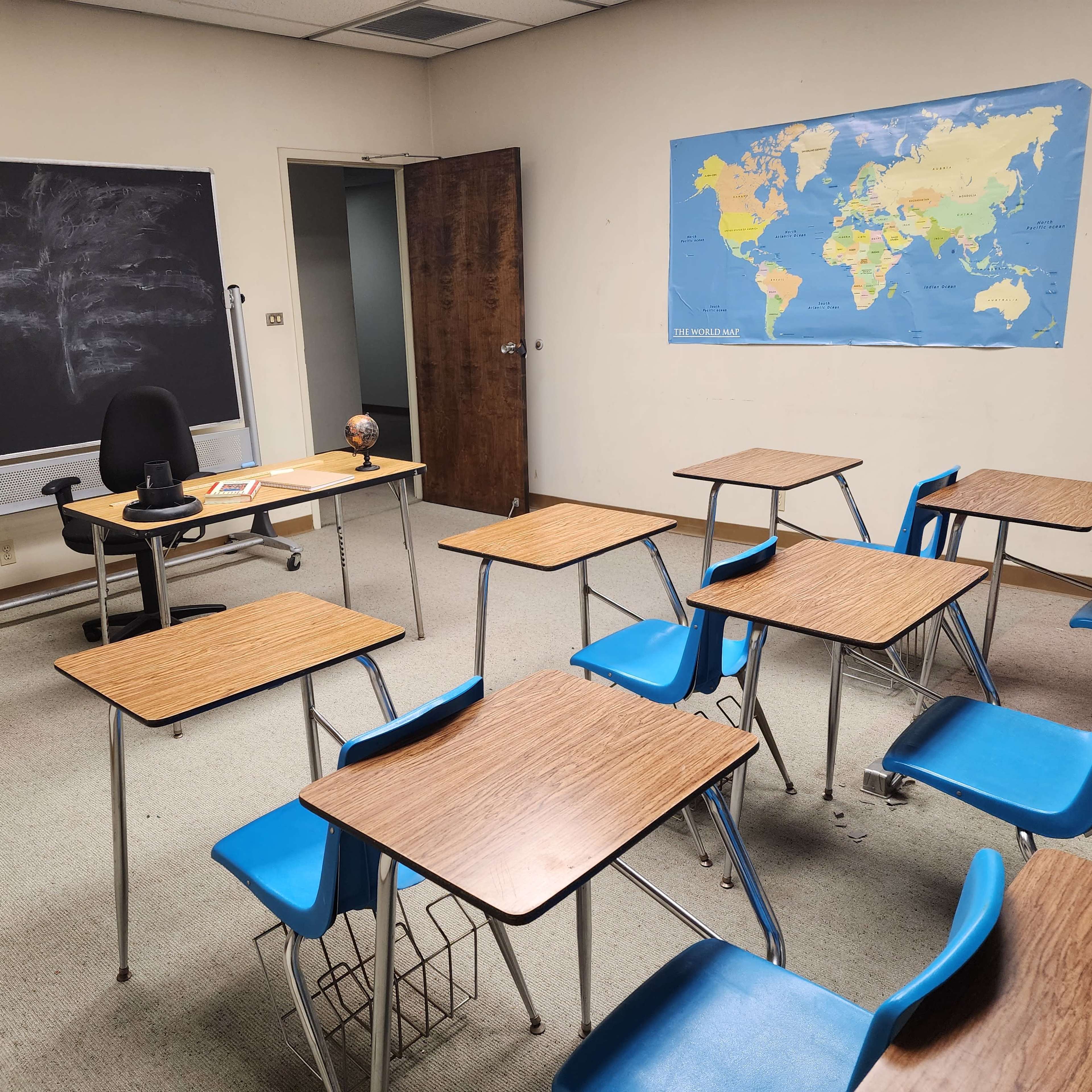A classroom is furnished with several empty desks, a chair, a globe, and a chalkboard, alongside a world map on the wall.