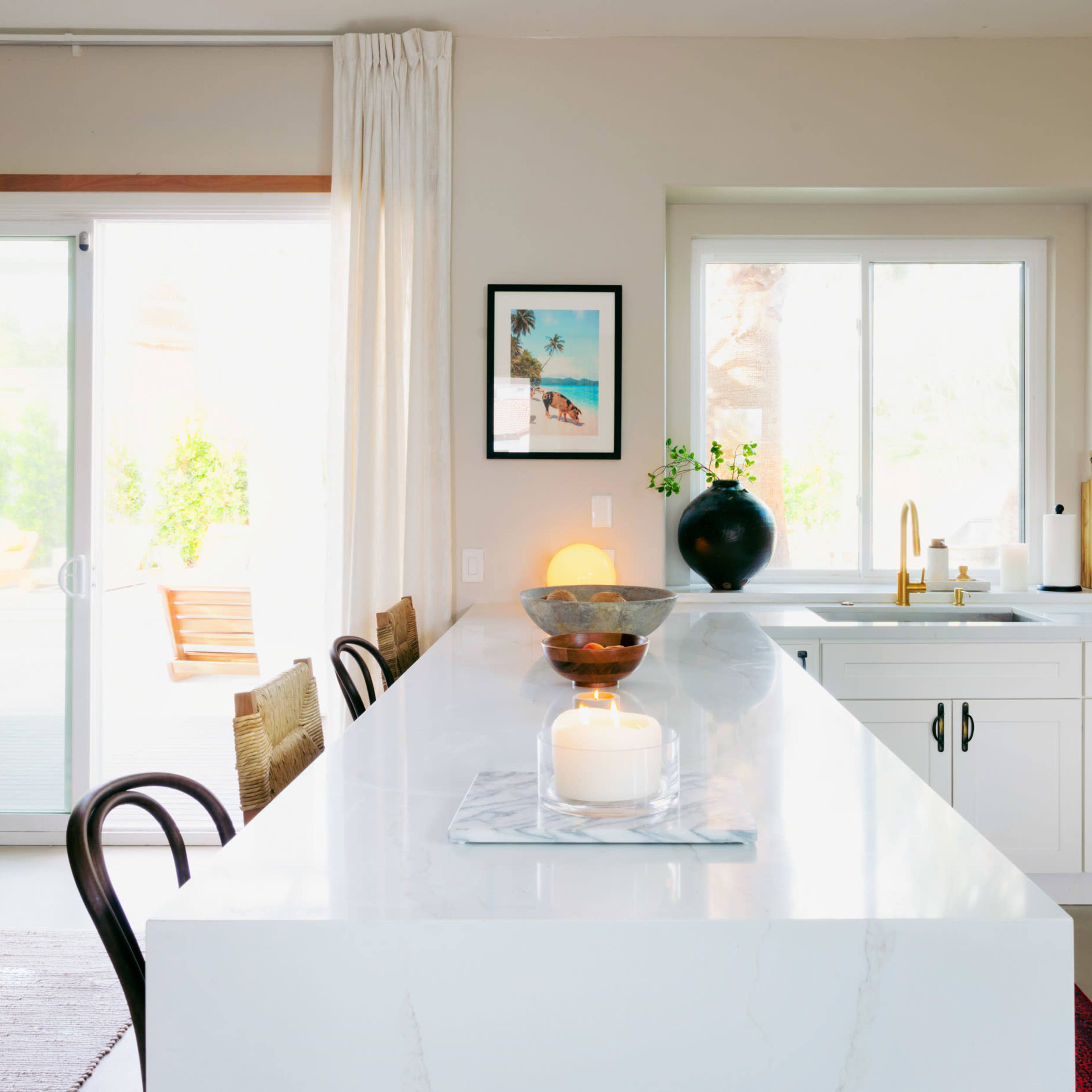 A modern kitchen features a large island with a marble countertop, white cabinets, and a window overlooking a patio area.