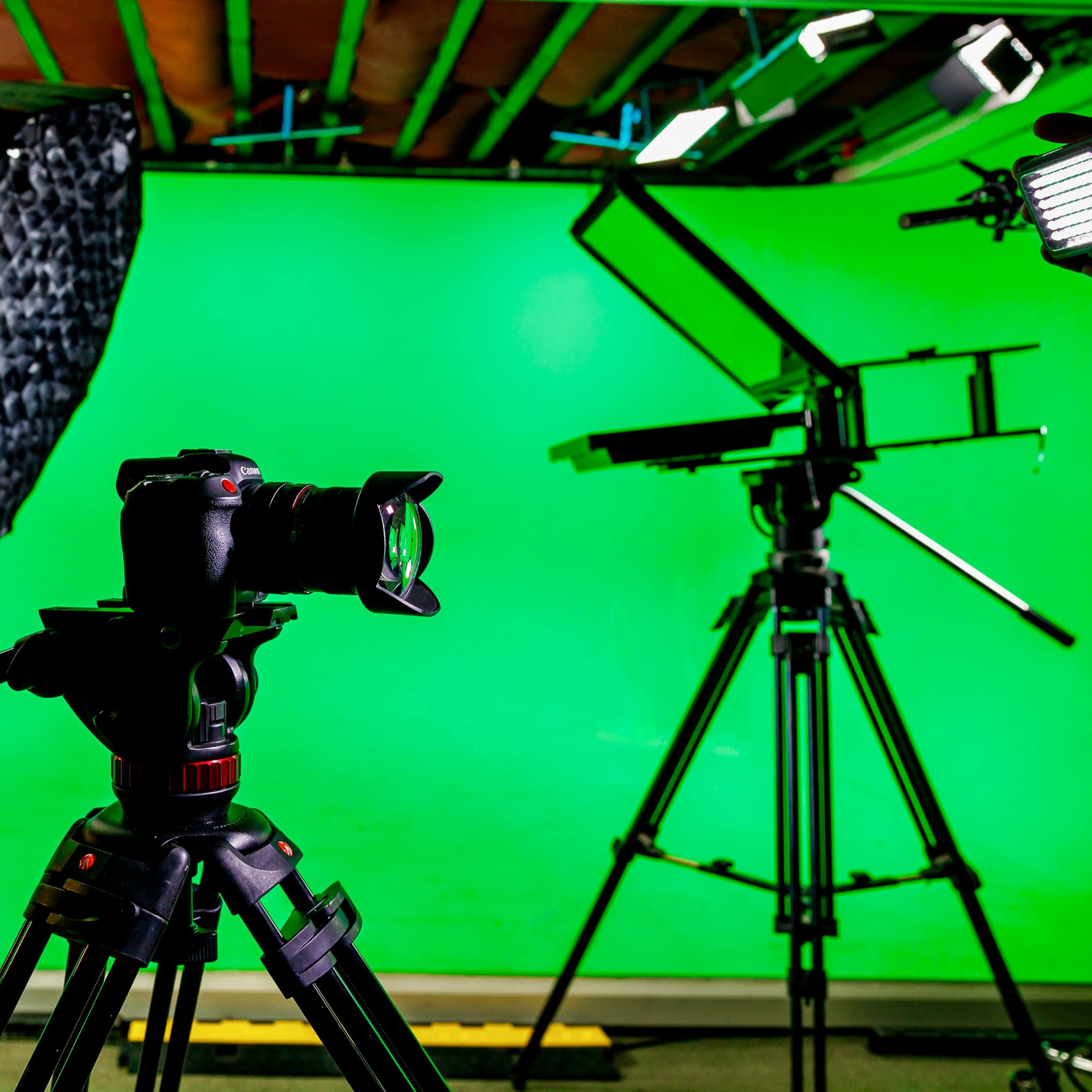 A camera is mounted on a tripod in front of a green screen setup, surrounded by studio lighting equipment.