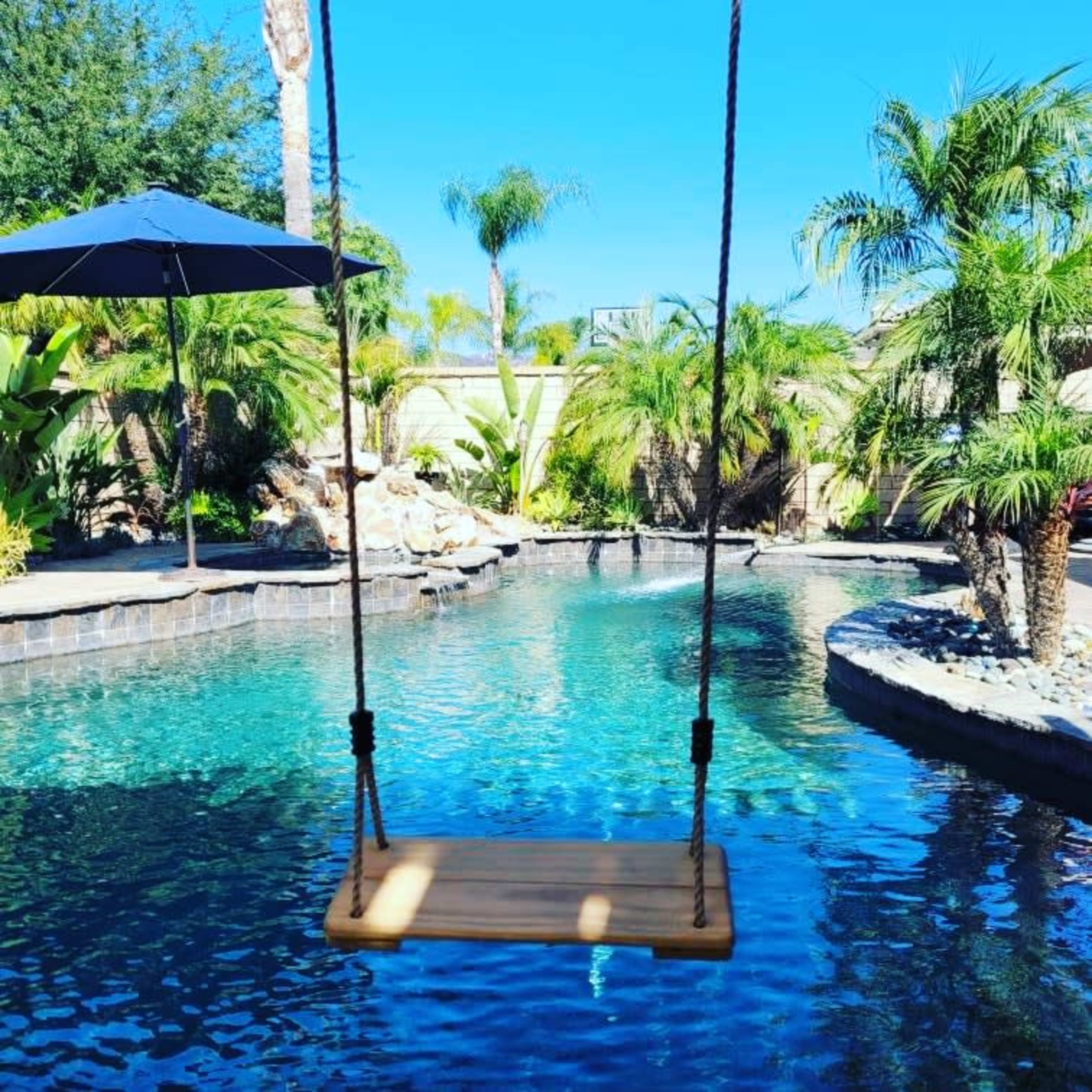 A wooden swing hangs over a clear blue pool surrounded by palm trees and lush greenery.