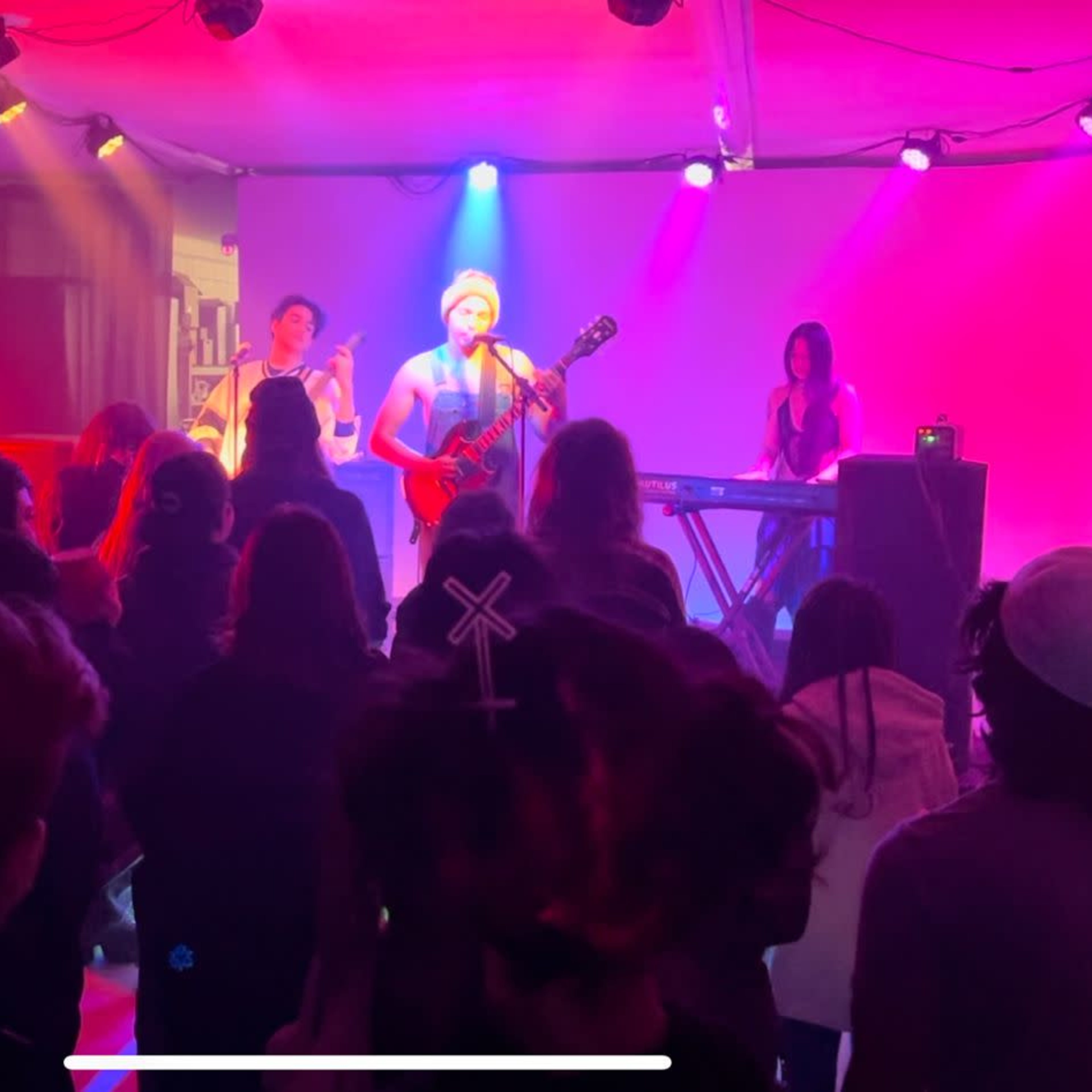 A band performs on stage in front of an audience in a dimly lit venue with colorful lighting.