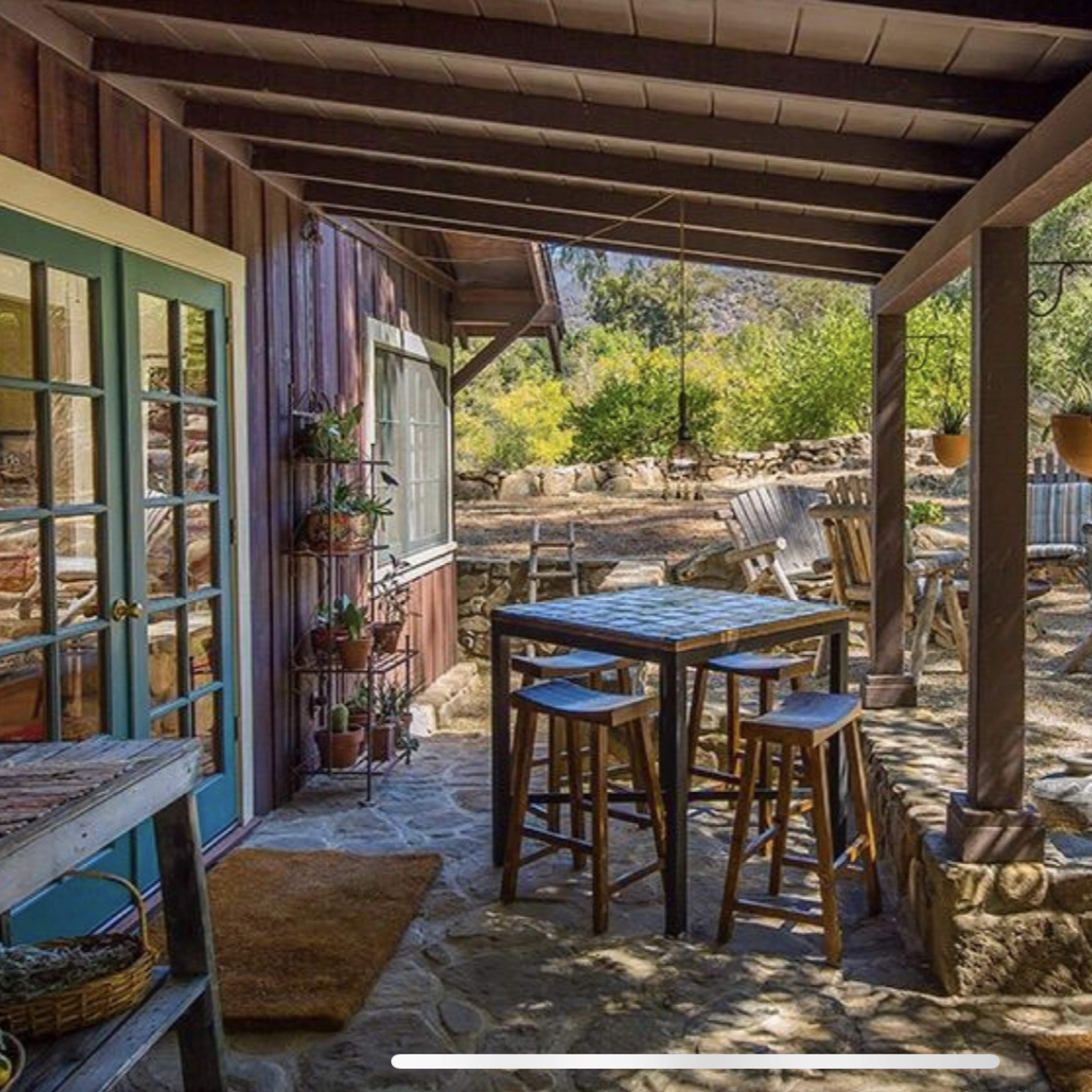 A rustic outdoor seating area features a wooden table and stools under a porch, surrounded by greenery and a stone pathway.