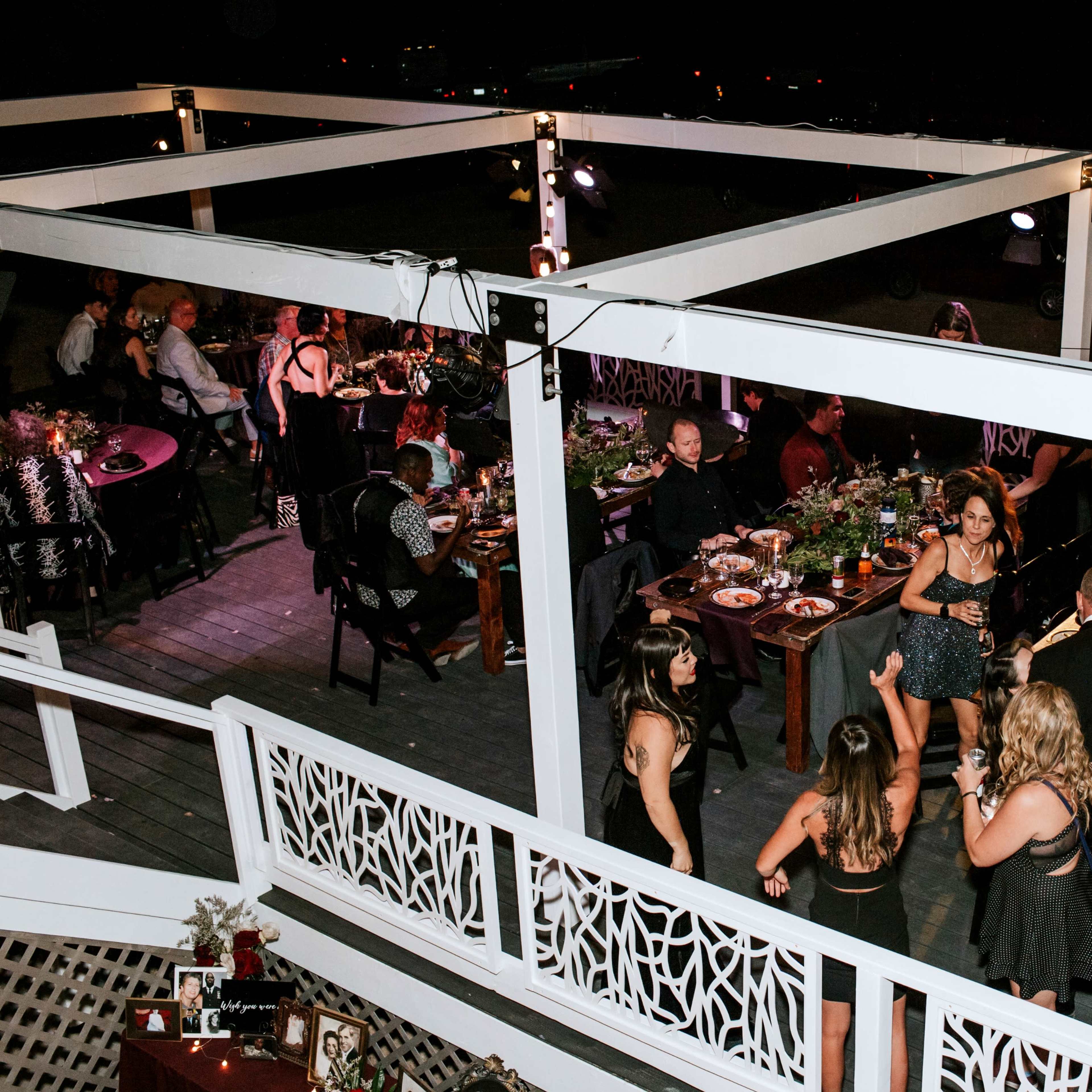 A group of people socialize and dine at a decorated outdoor event with tables set under decorative lighting.