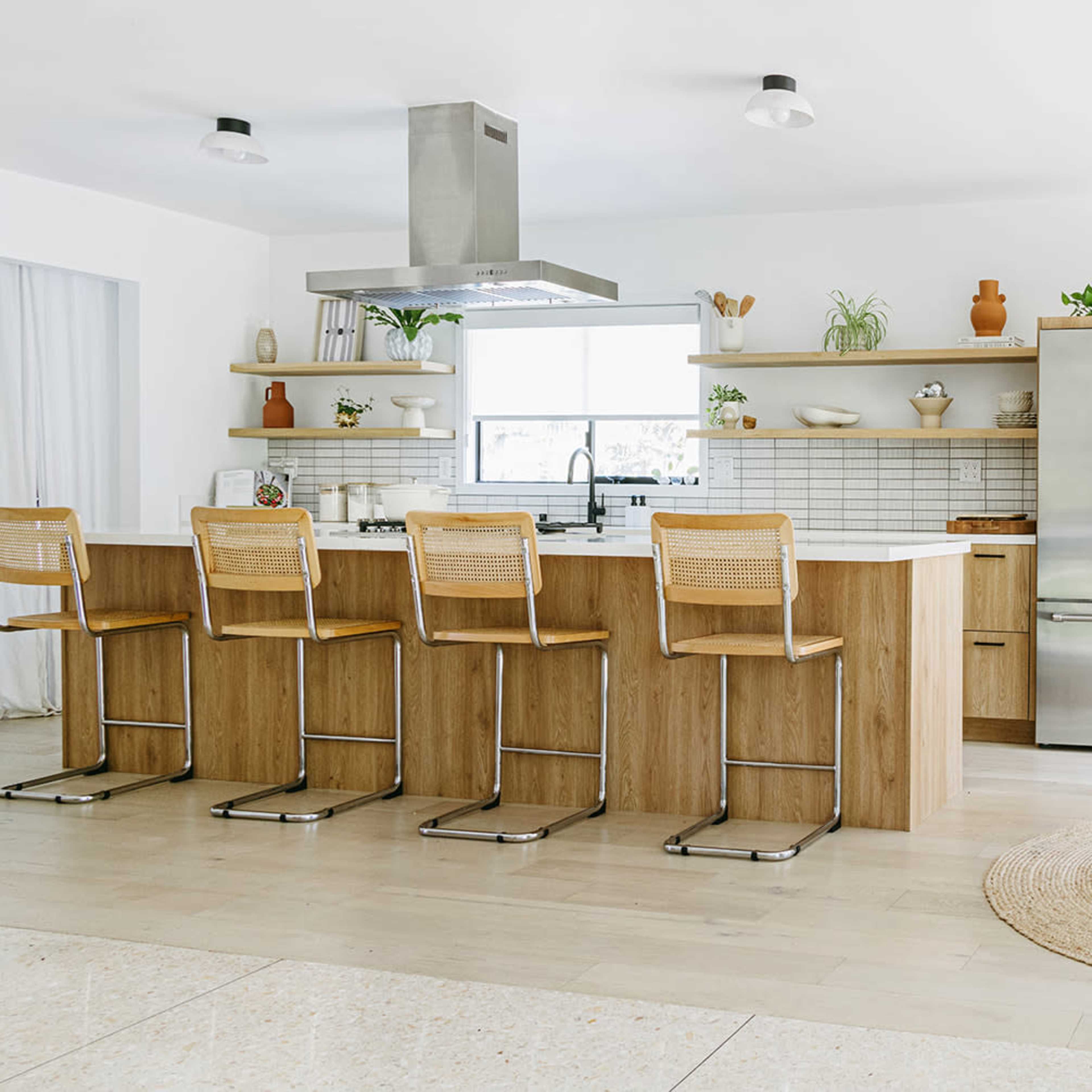 A modern kitchen features a large island with four bar stools, stainless steel appliances, and shelves adorned with plants and dishware.