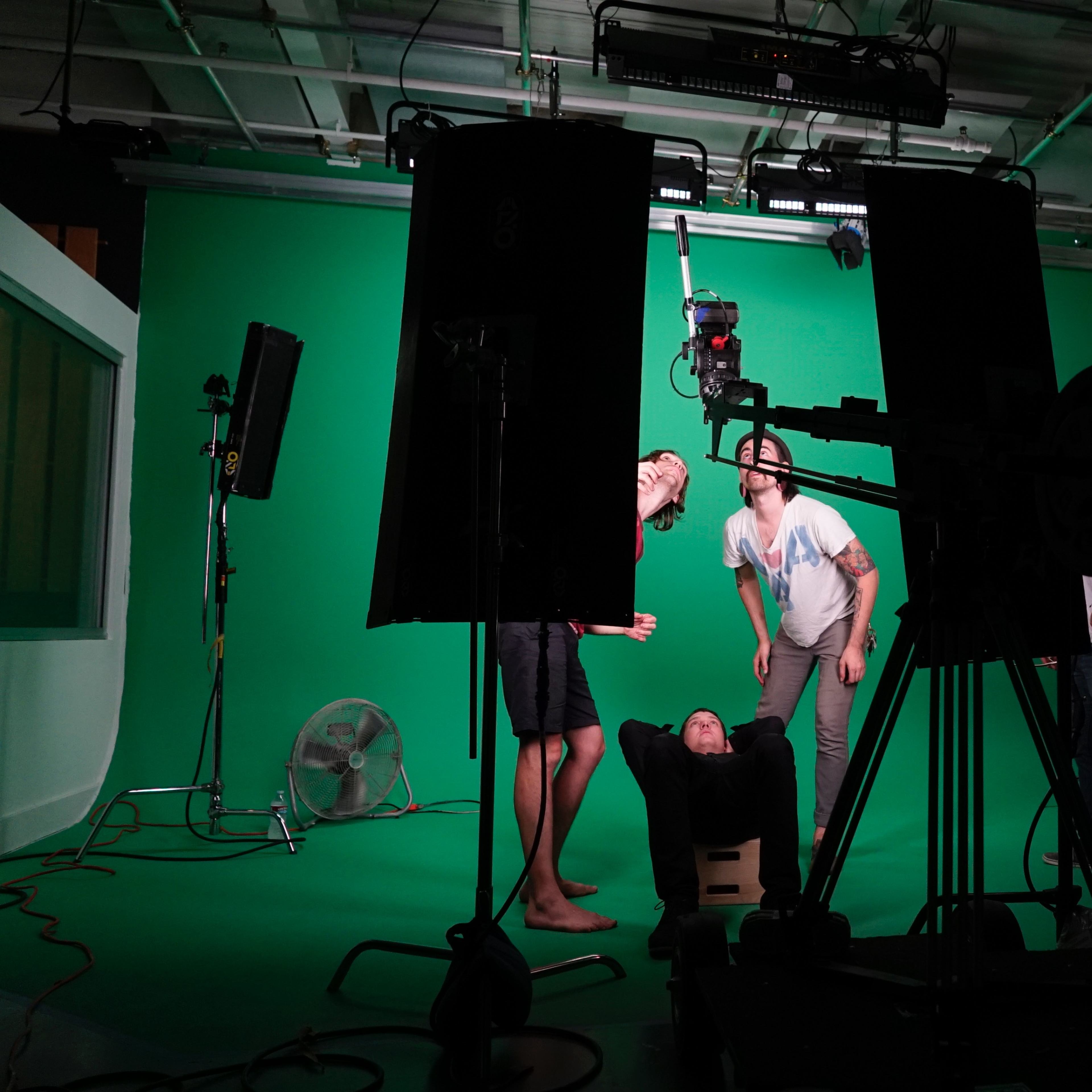 A group of people are positioned in a studio with a green screen, with one individual lying on a cart while others interact around them, and various filming equipment is visible in the foreground.