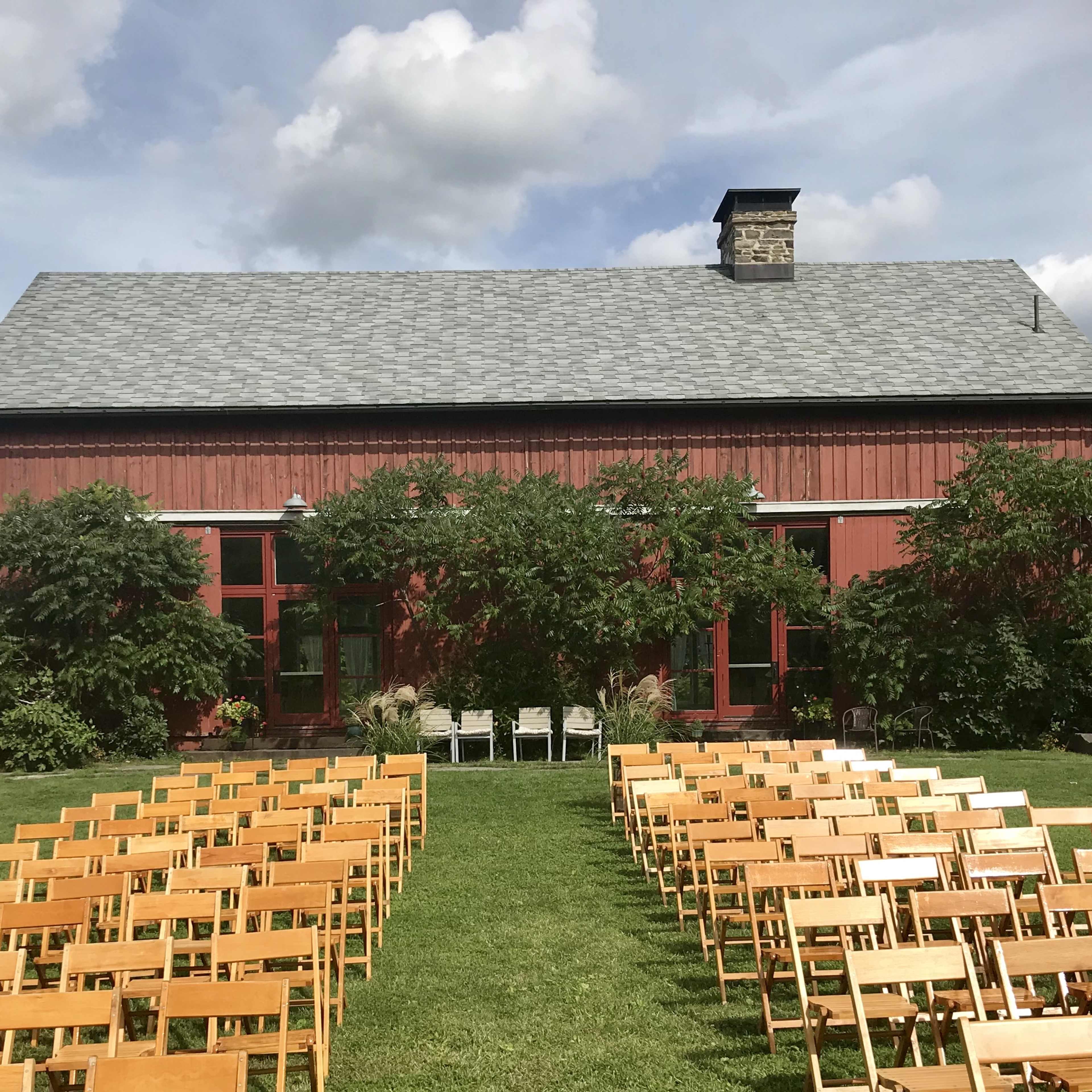The scene shows a garden setting with rows of wooden chairs arranged on a grassy area in front of a red barn.