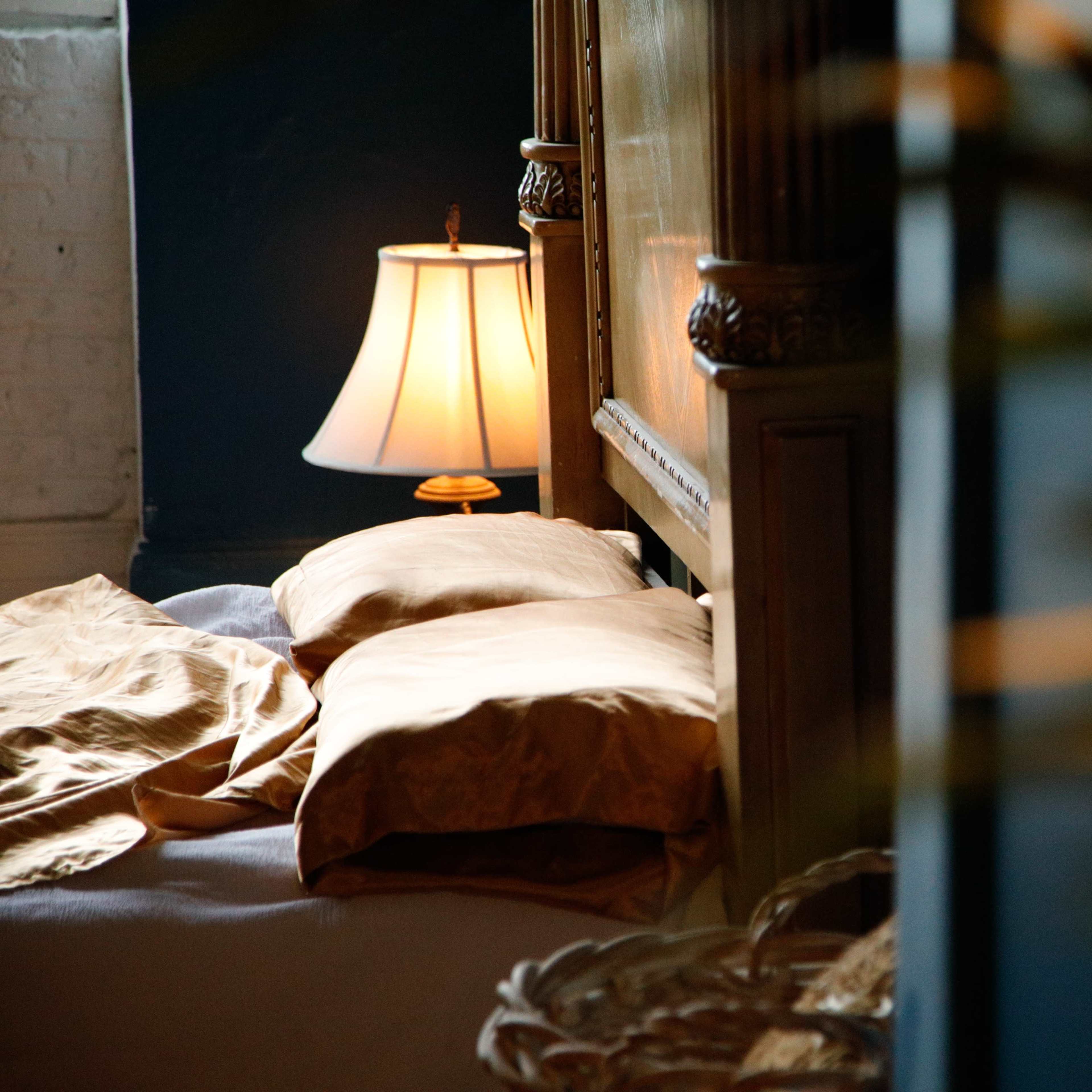 A softly lit bedroom features a bed with two beige pillows, partially covered by a light-colored blanket, and a bedside lamp casting a warm glow.