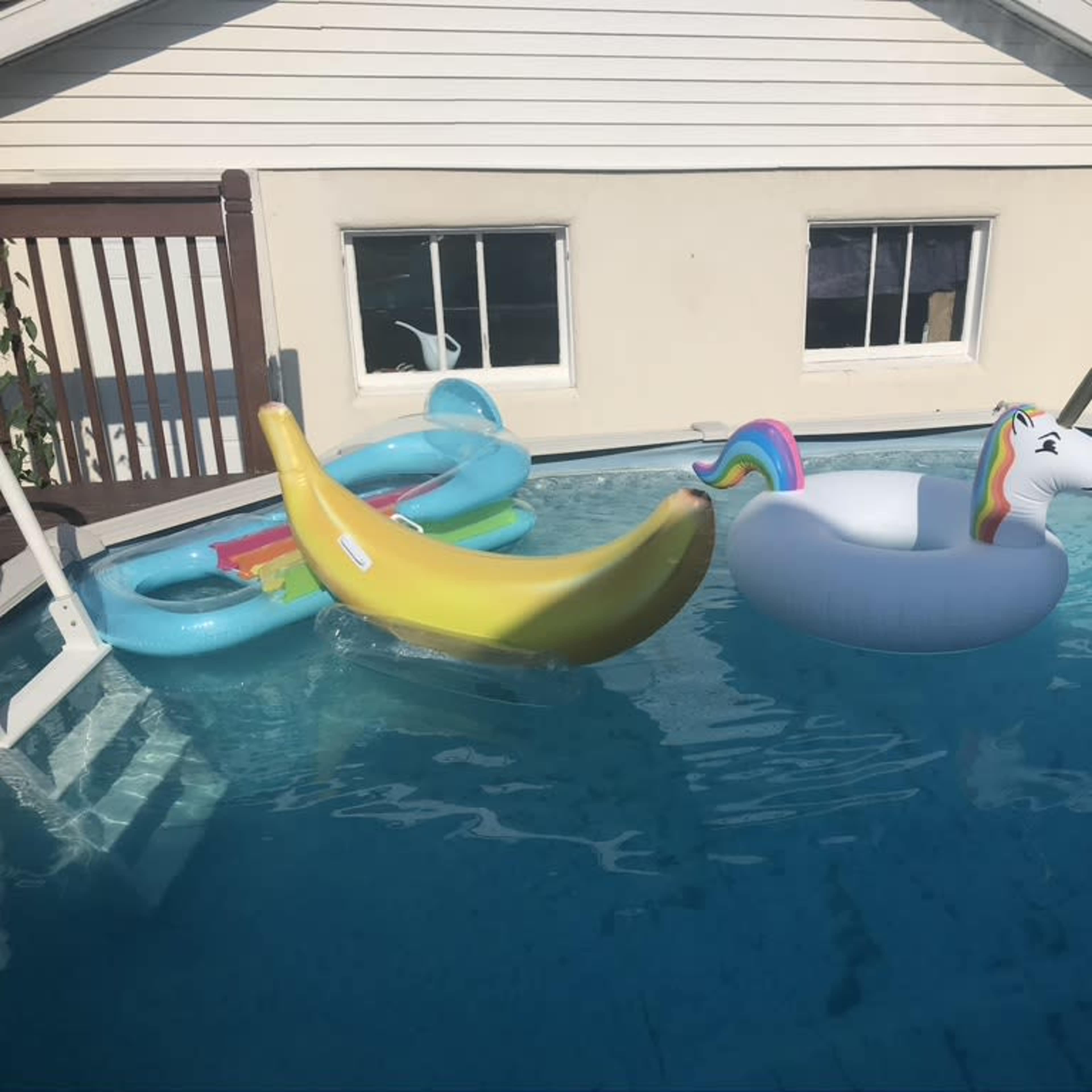 A unicorn-shaped float and a banana-shaped float are partially submerged in a swimming pool beside a house.