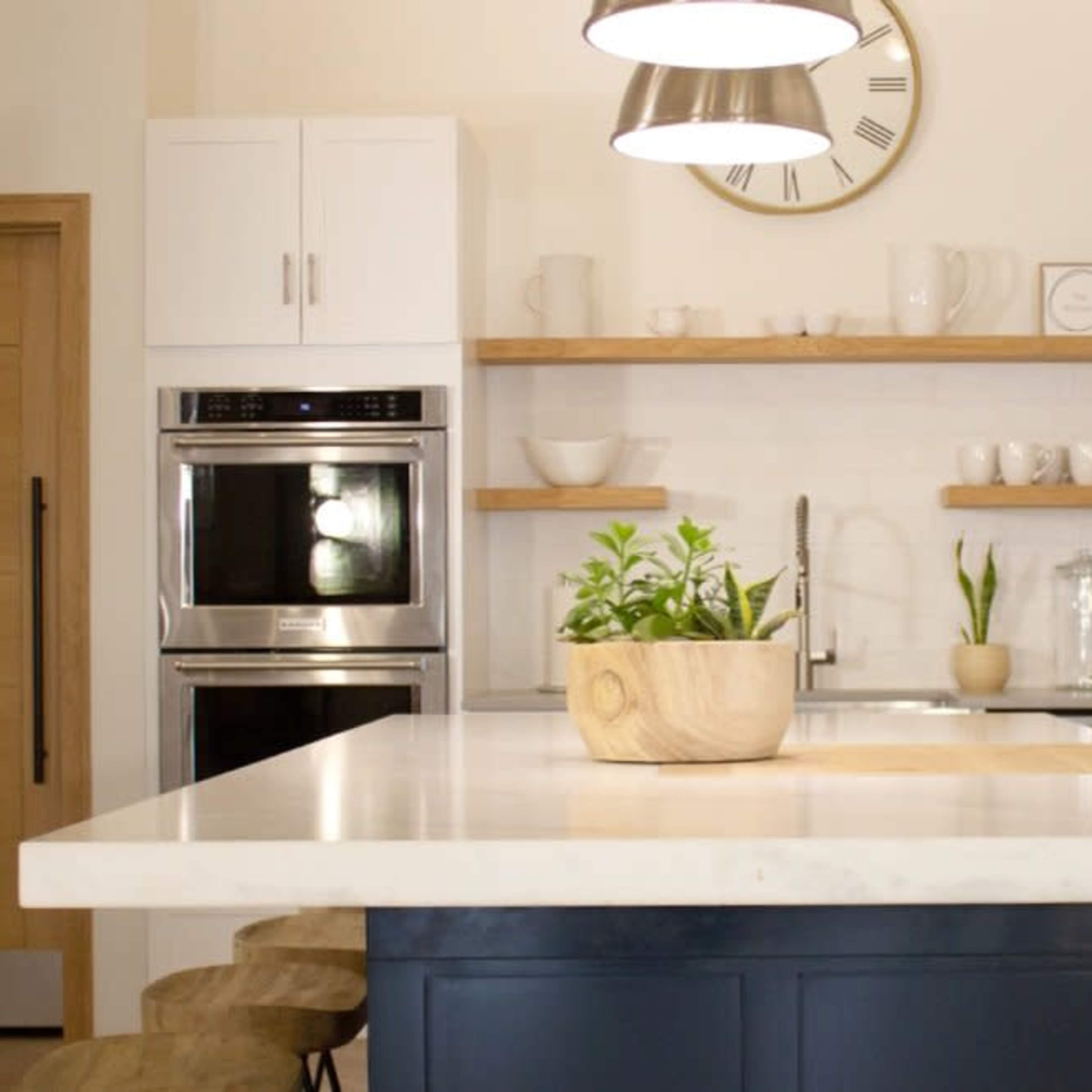 A modern kitchen features a central island with a plant in a wooden bowl, stainless steel appliances, and open shelves displaying dishware.