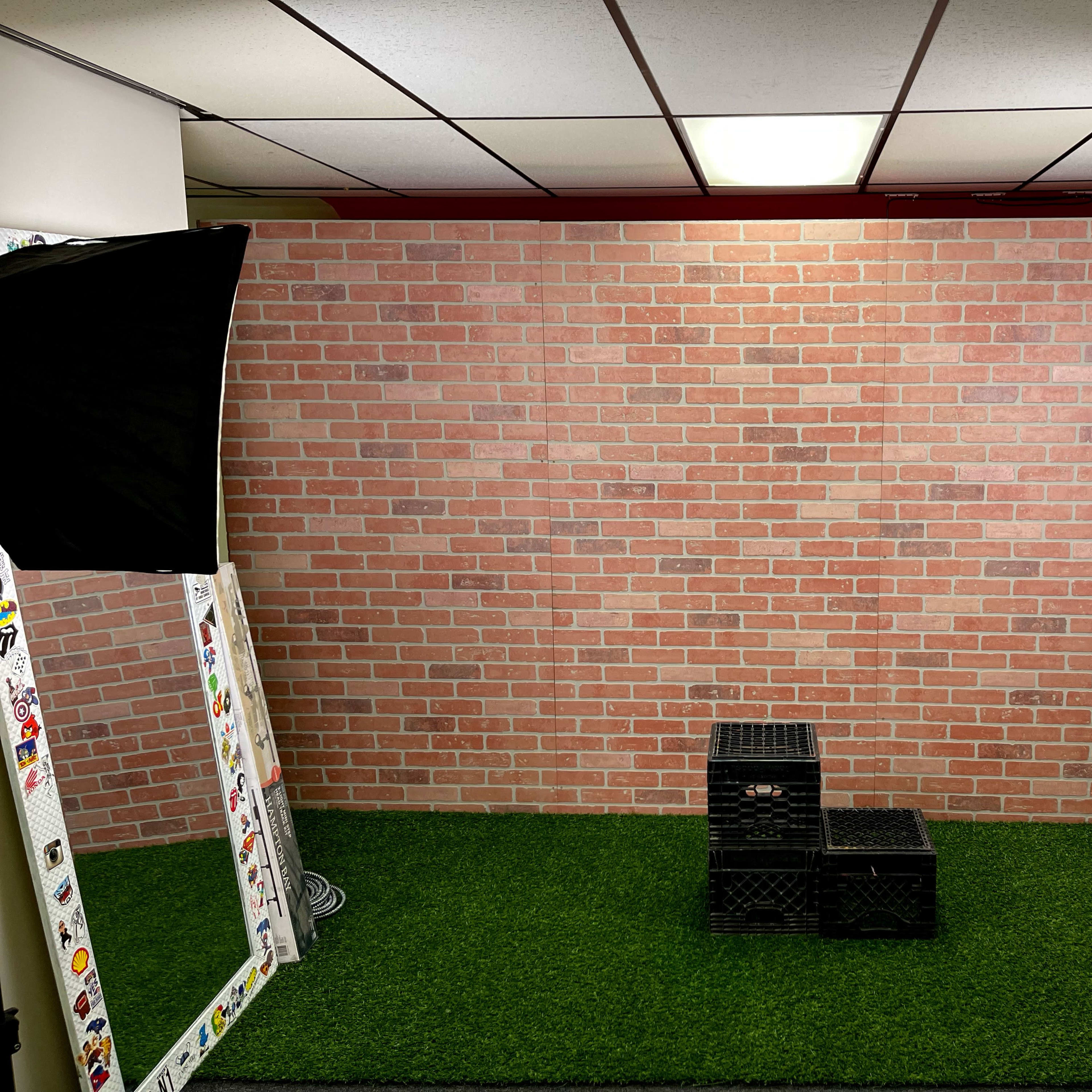 A studio space featuring a brick wall backdrop, two lighting setups, a mirror, and black crates on a green turf floor.