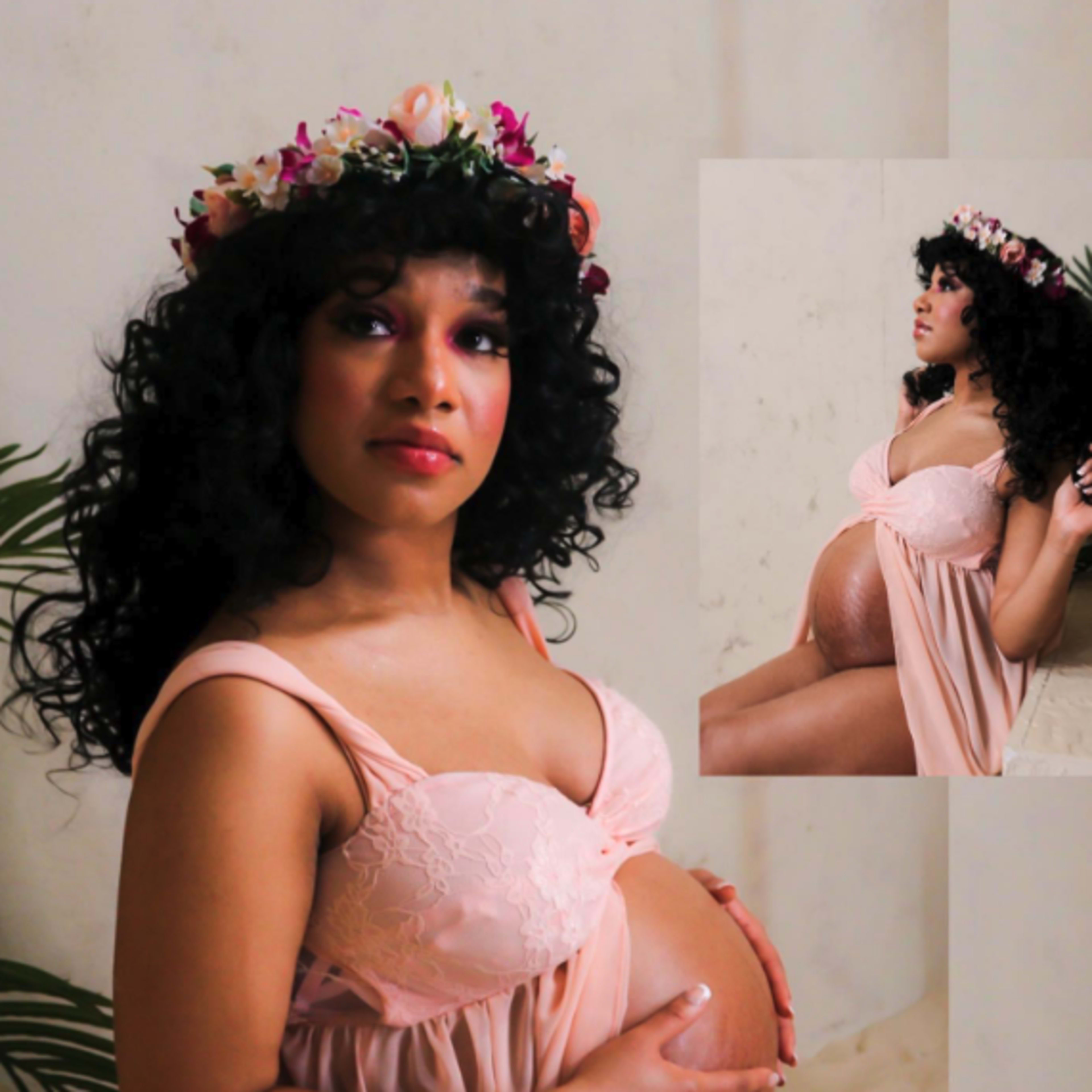 A pregnant woman in a pink dress with a floral crown sits gracefully against a plain backdrop, gently cradling her belly.
