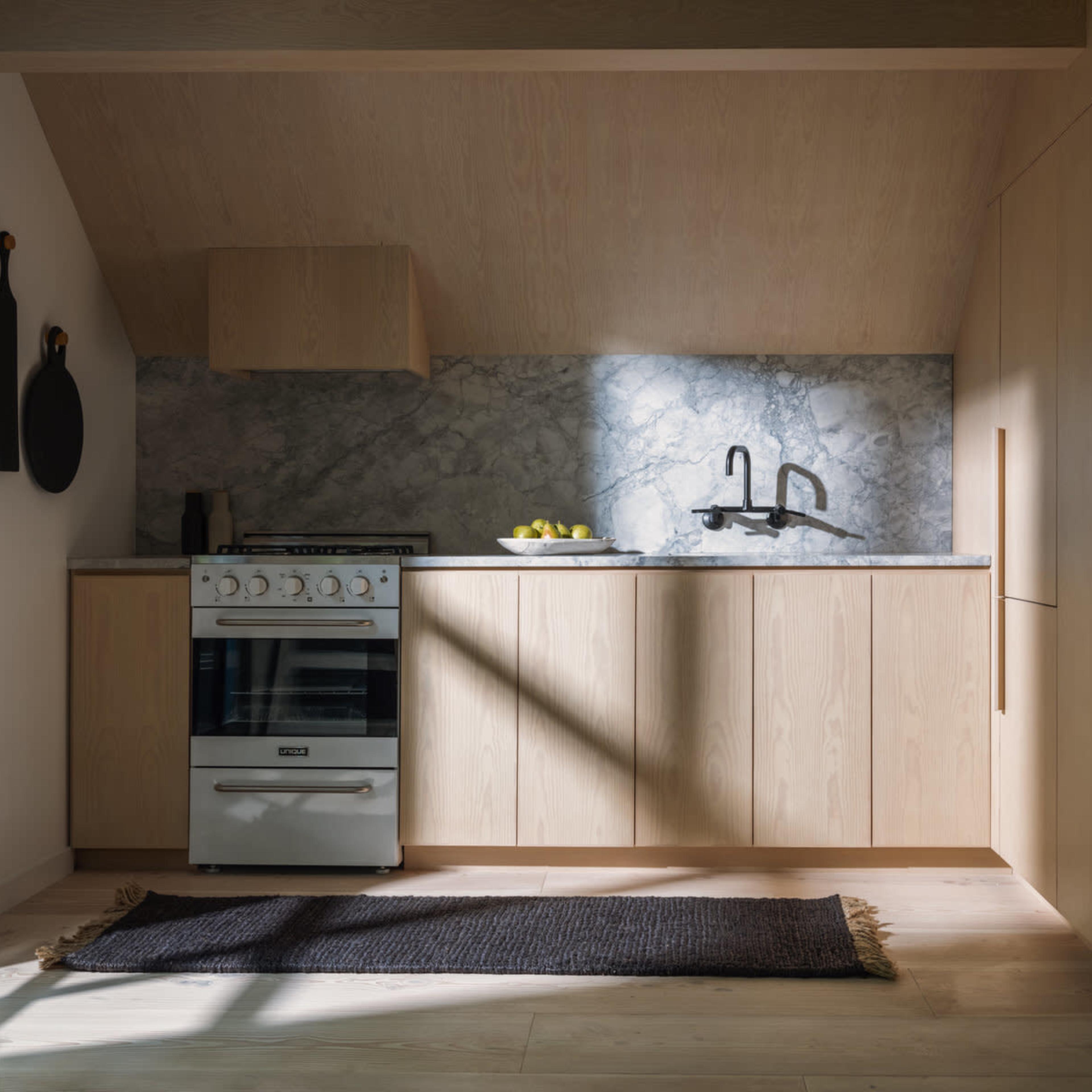A modern kitchen features light wood cabinetry, a marble backsplash, a stainless steel stove, and a dark rug on the floor.