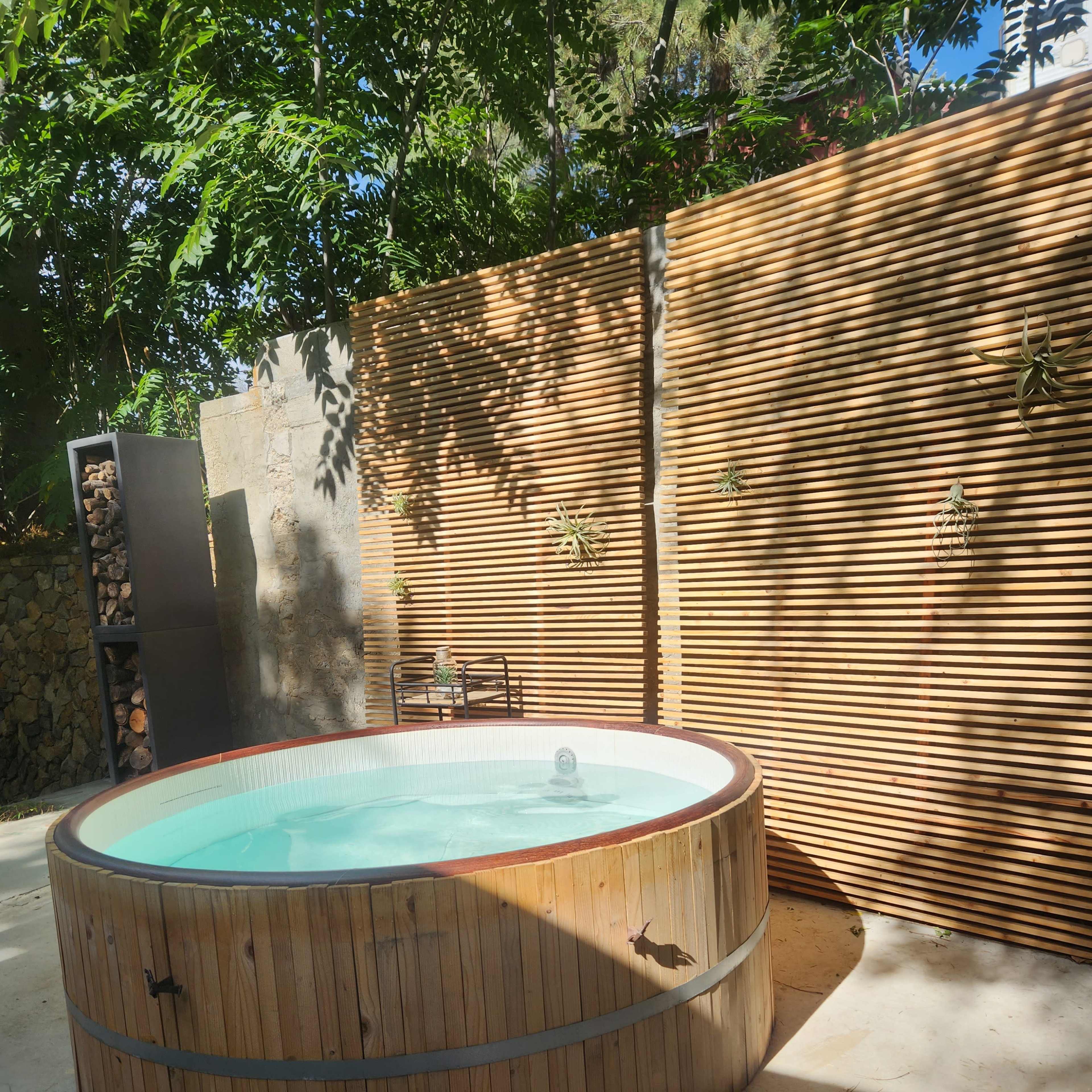 A round wooden hot tub surrounded by a wooden slatted wall and greenery.