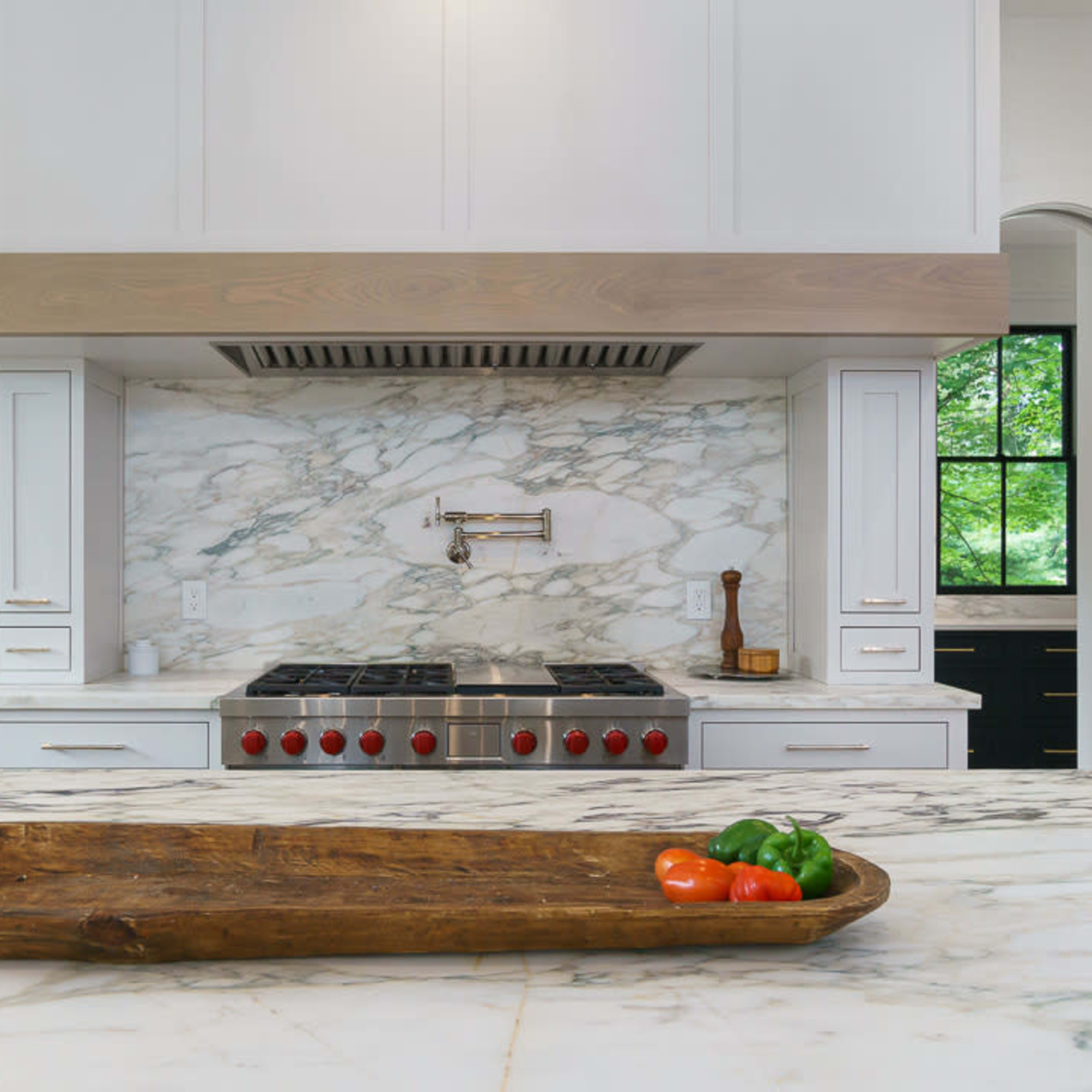 A modern kitchen featuring a marble backsplash, a professional-grade stovetop with red knobs, and a wooden tray with tomatoes and bell peppers on a marble countertop.