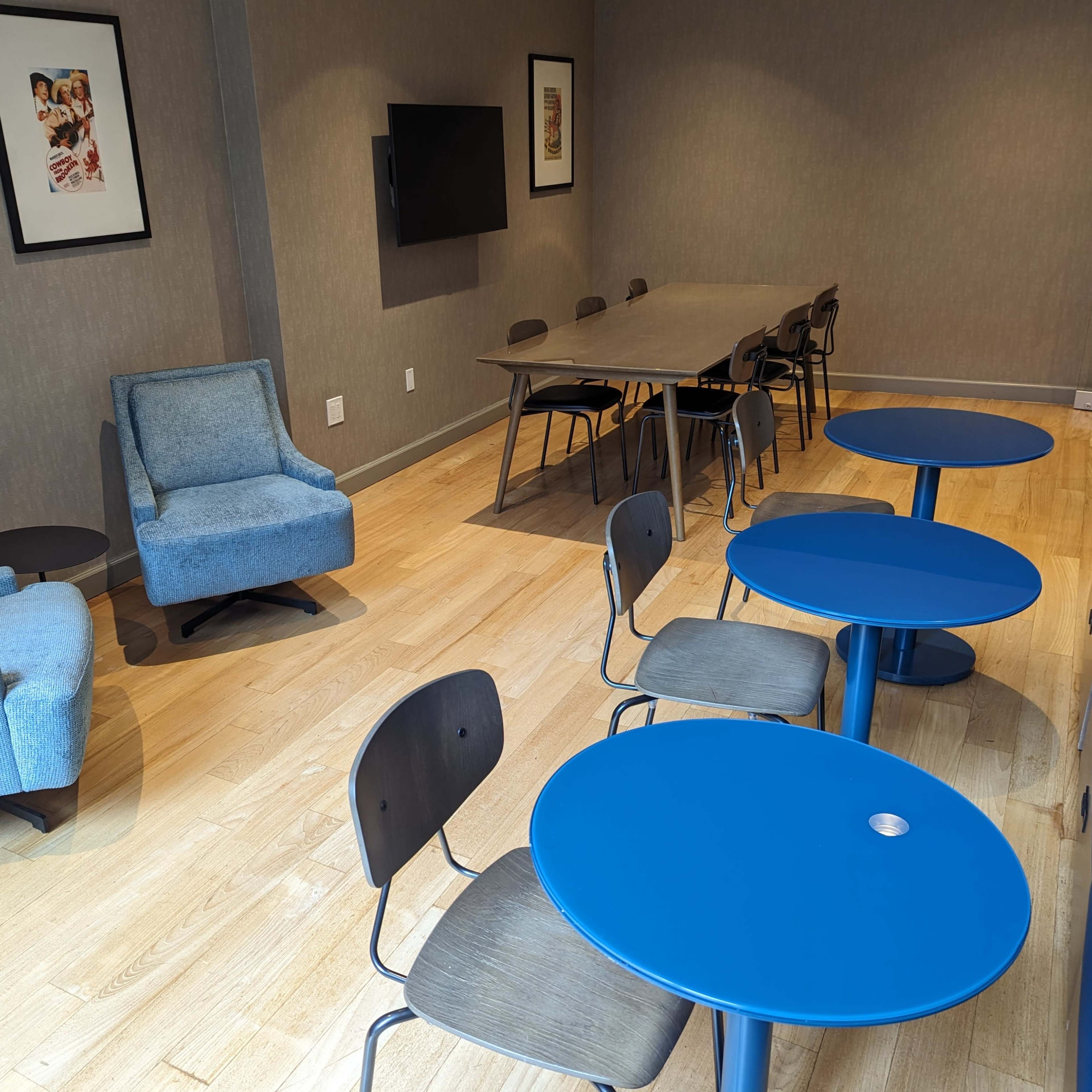 A well-lit room featuring two blue armchairs, several blue tables, black chairs, a large table, and framed artwork on the walls.