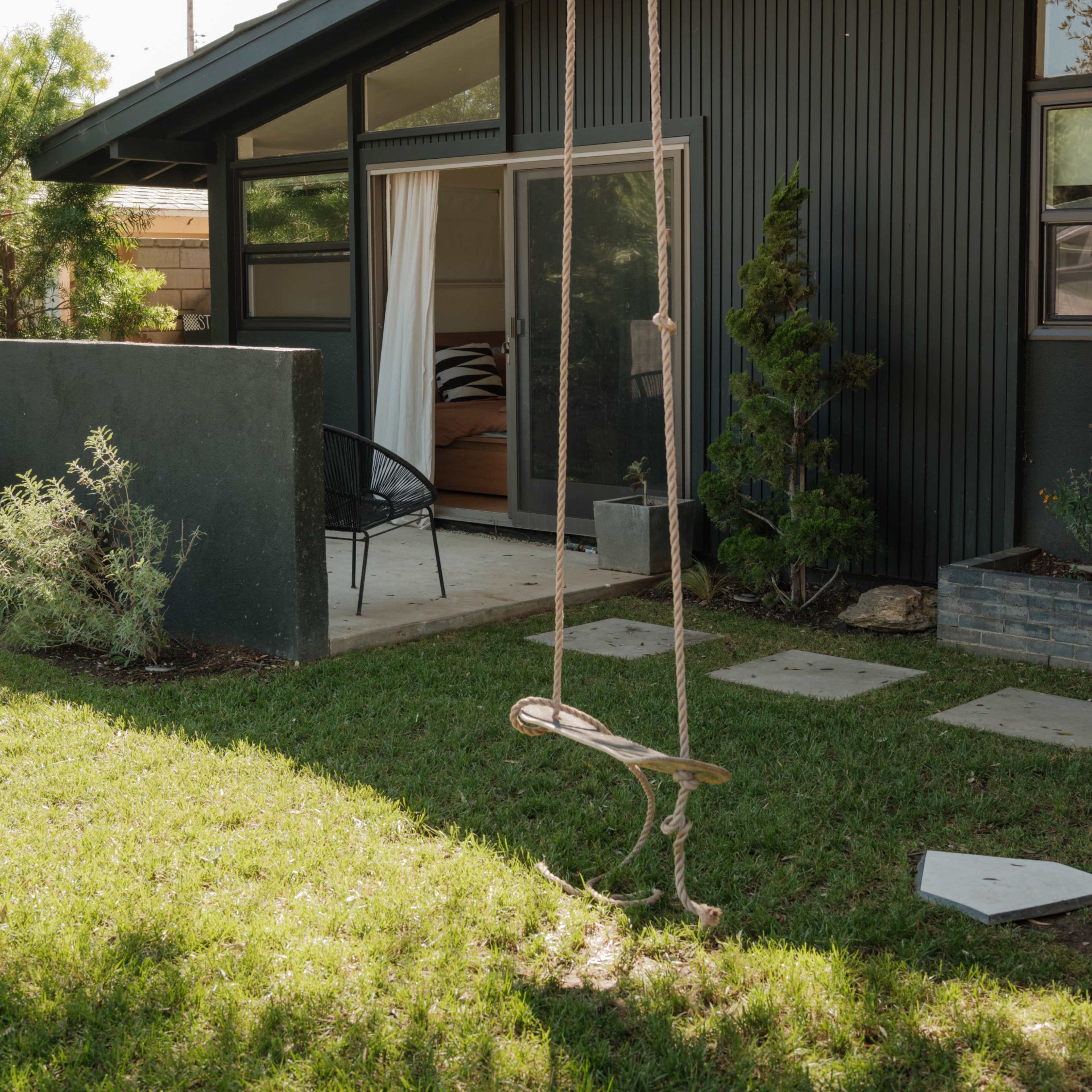 A swing hangs from a tree in a grassy yard beside a modern, dark-colored house with large windows and a patio door.