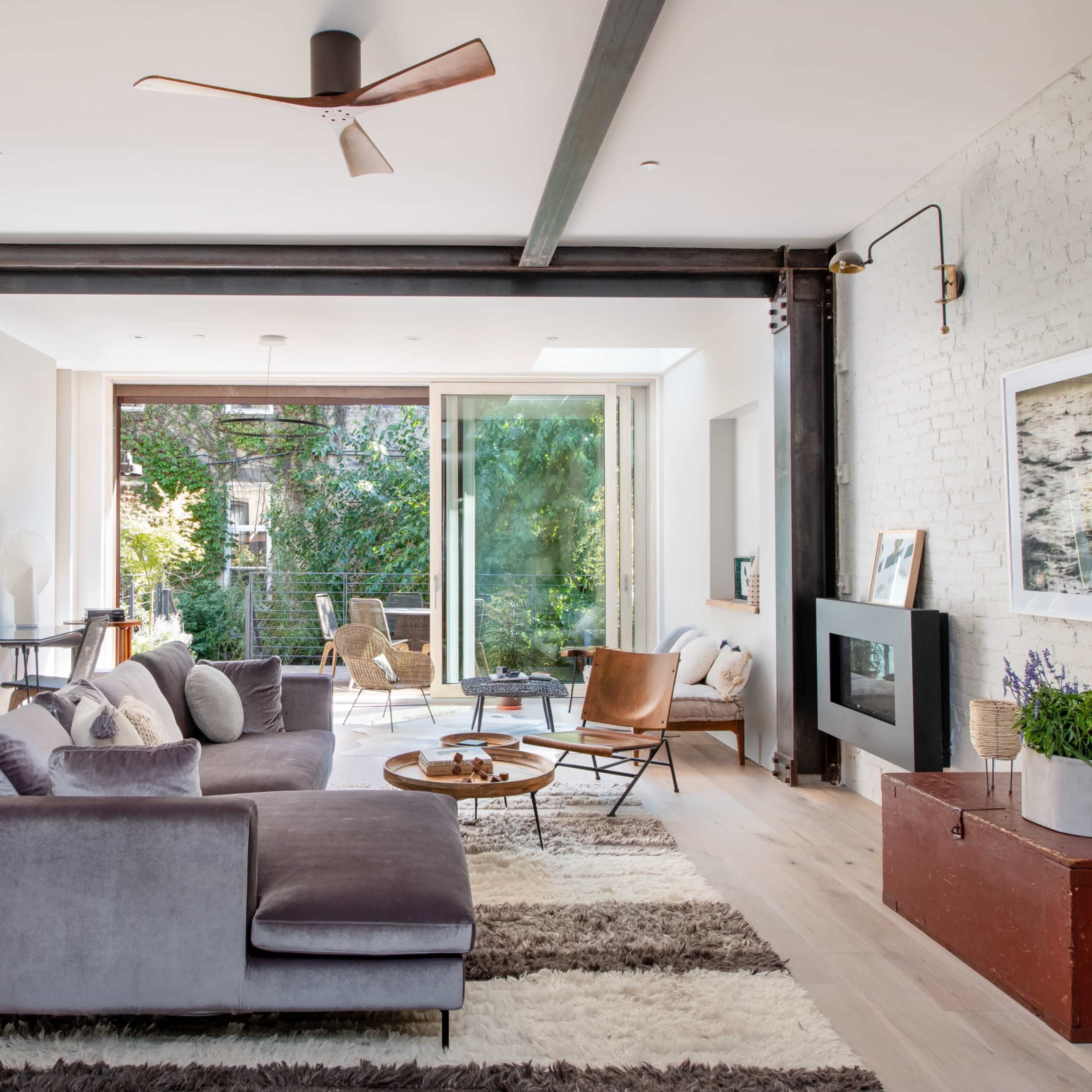 A modern living room features a gray sectional sofa, a wooden coffee table, and large windows that open to a balcony with greenery outside.