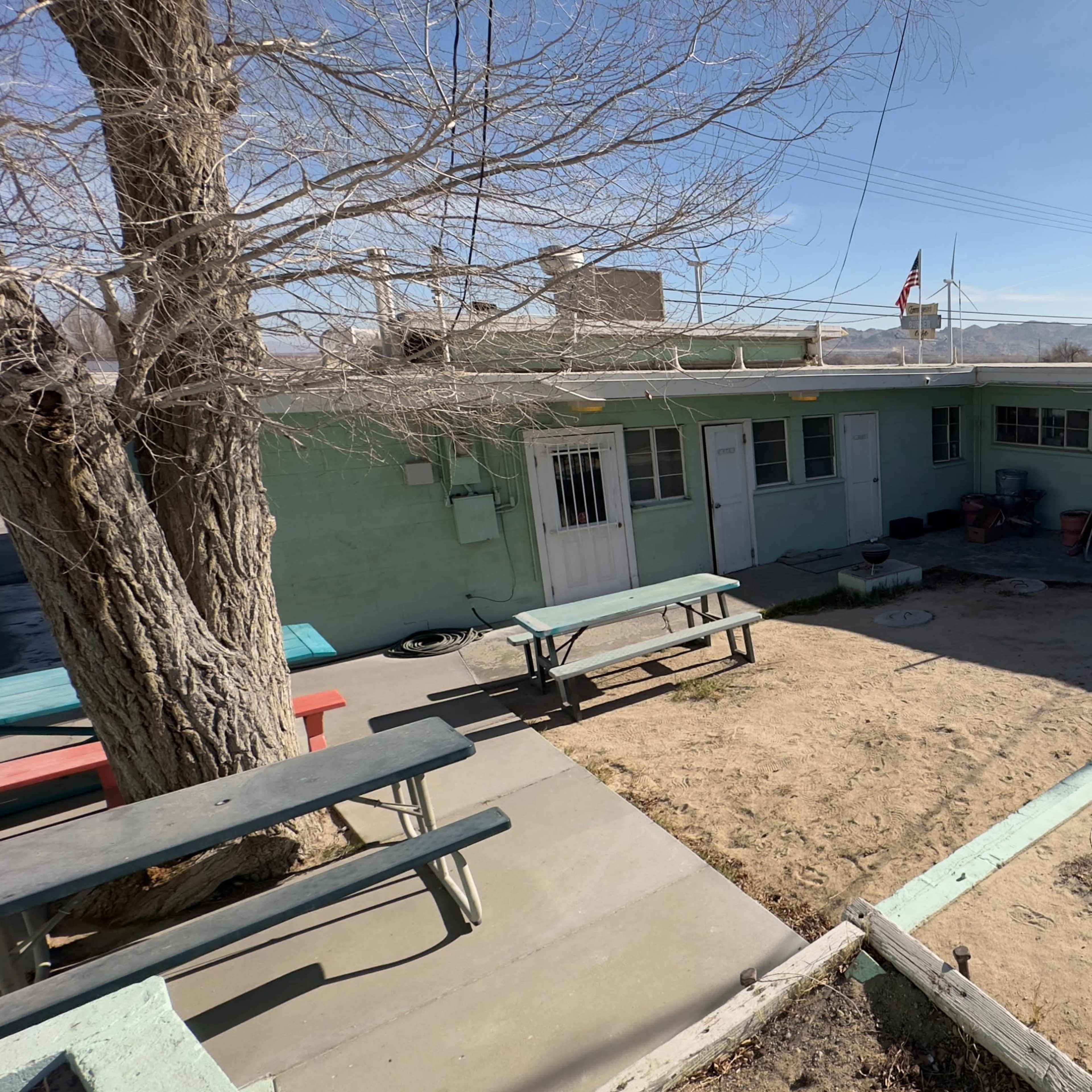 A single-story, light green building with a courtyard area, picnic tables, and a bare tree in the foreground under a clear blue sky.