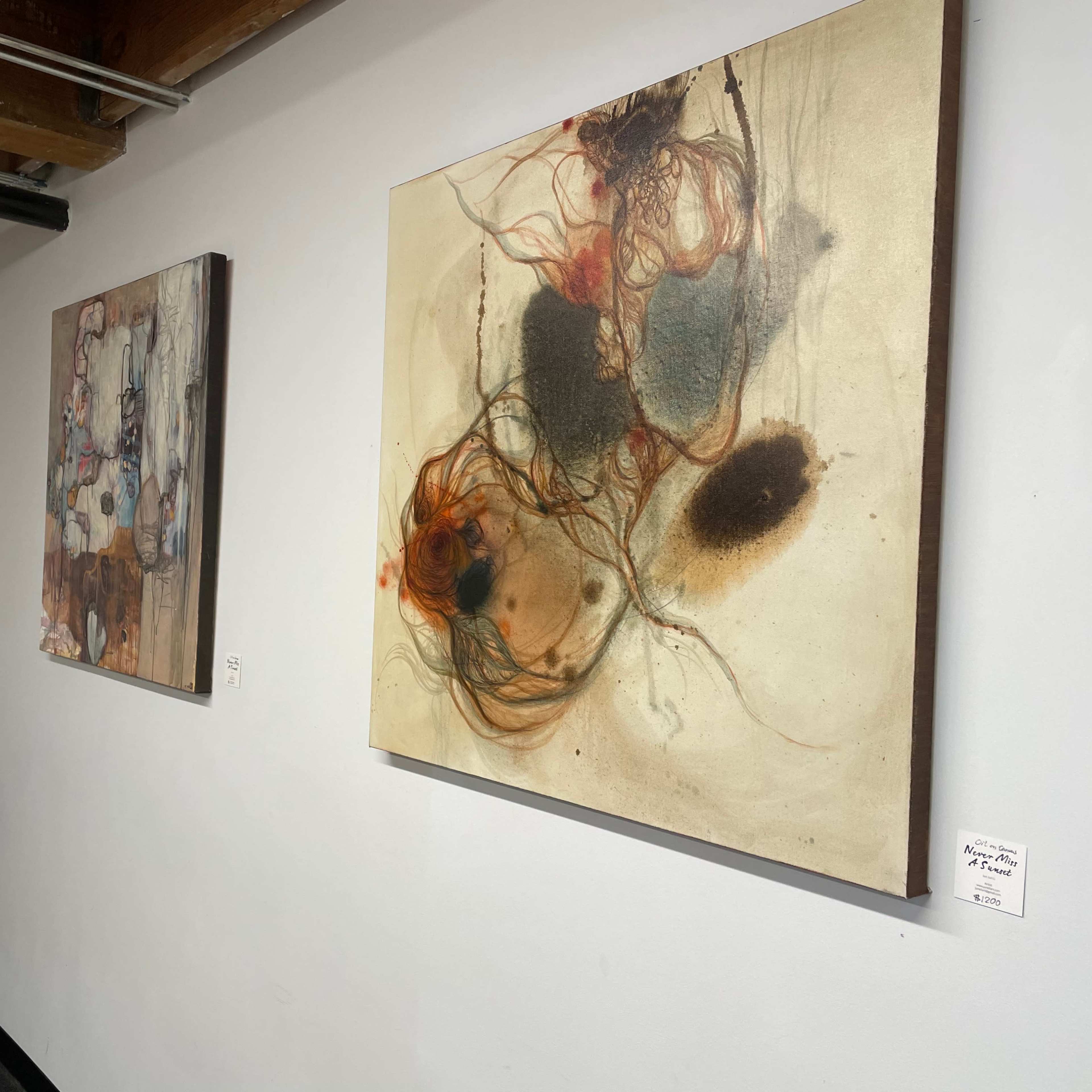 Two abstract paintings displayed on a white wall in an art gallery, with a metal stool positioned nearby.