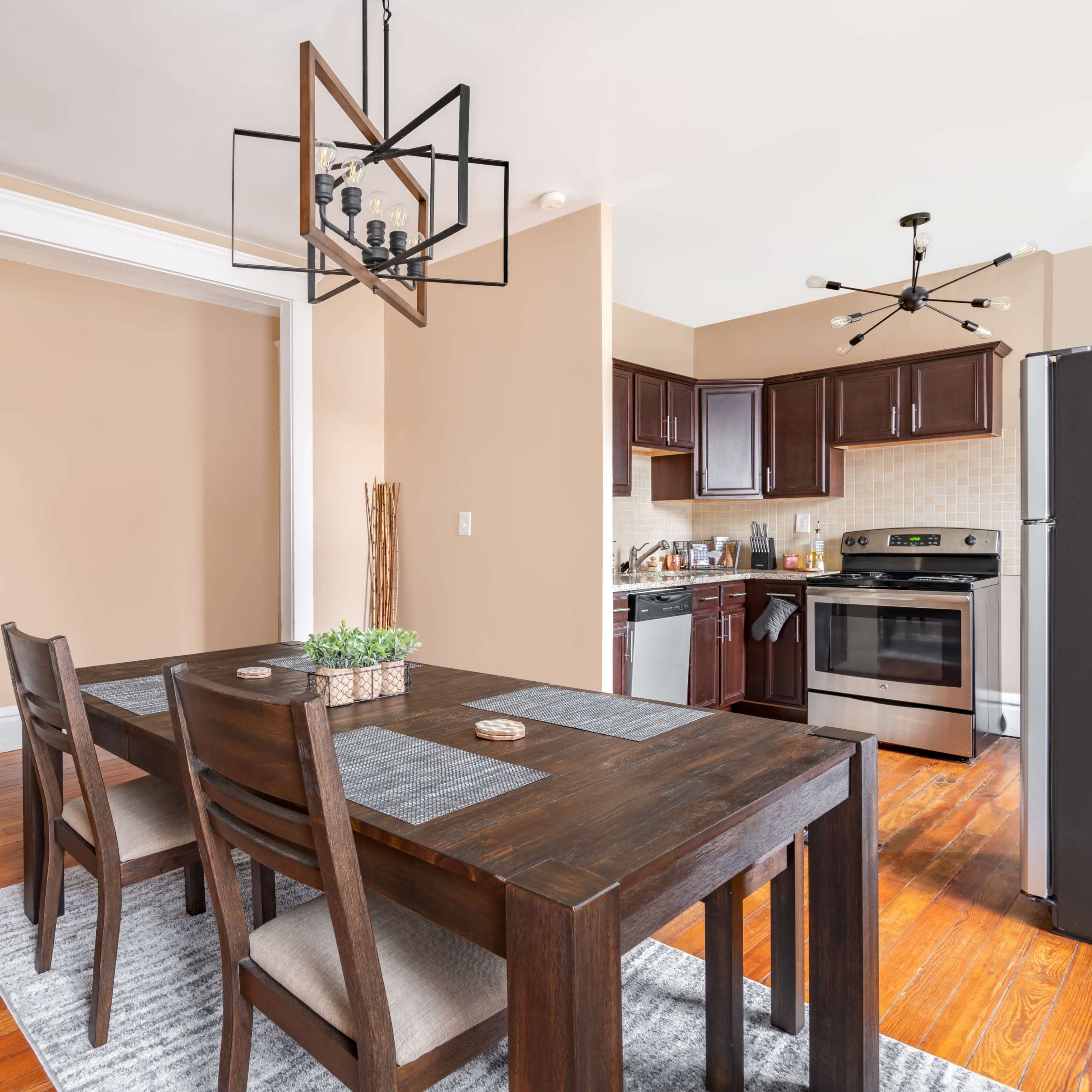 A modern dining area featuring a wooden table with chairs, a pendant light fixture above, and an adjacent kitchen with dark cabinetry and stainless steel appliances.