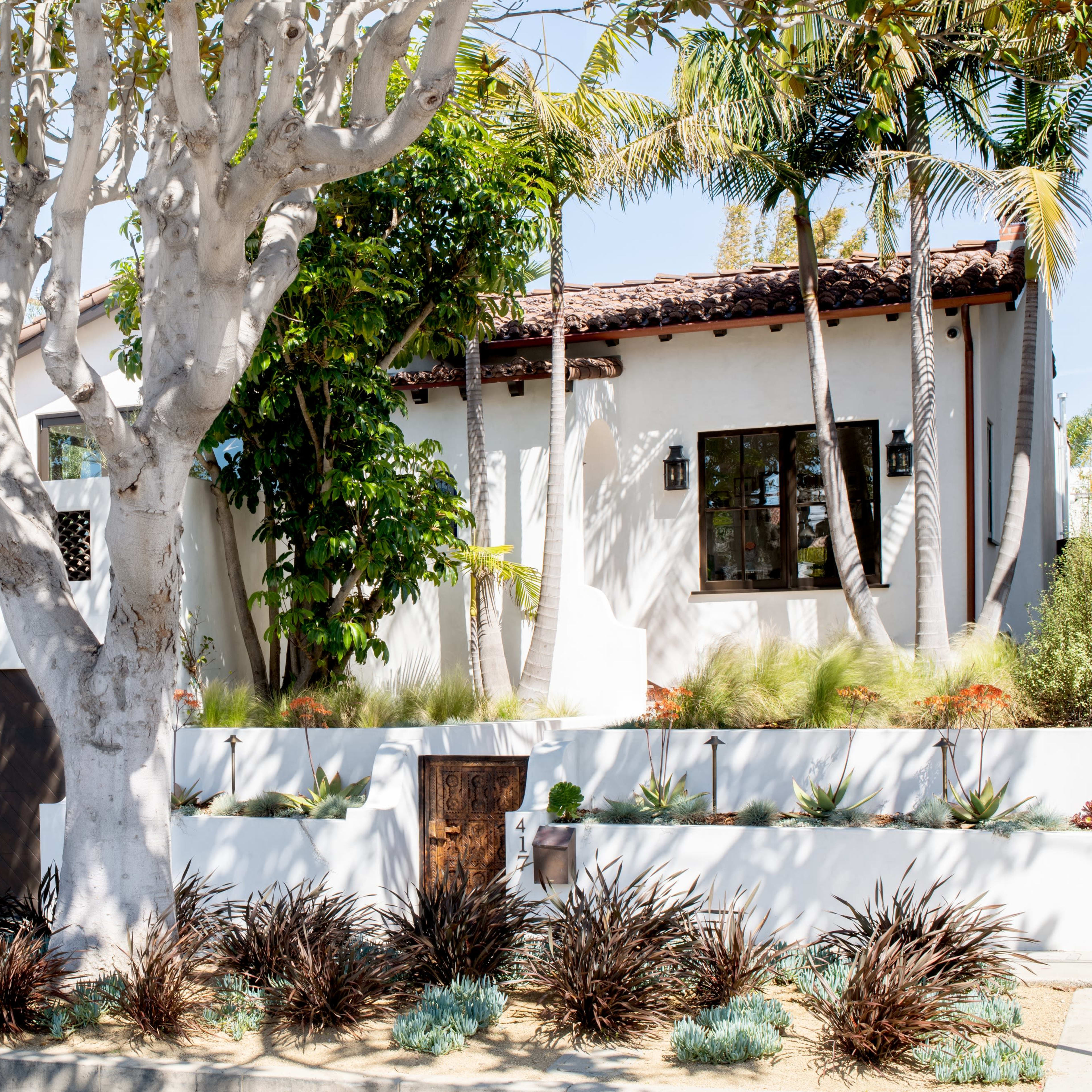 A white house with a dark garage door, surrounded by palm trees and landscaping, sits behind a low wall.