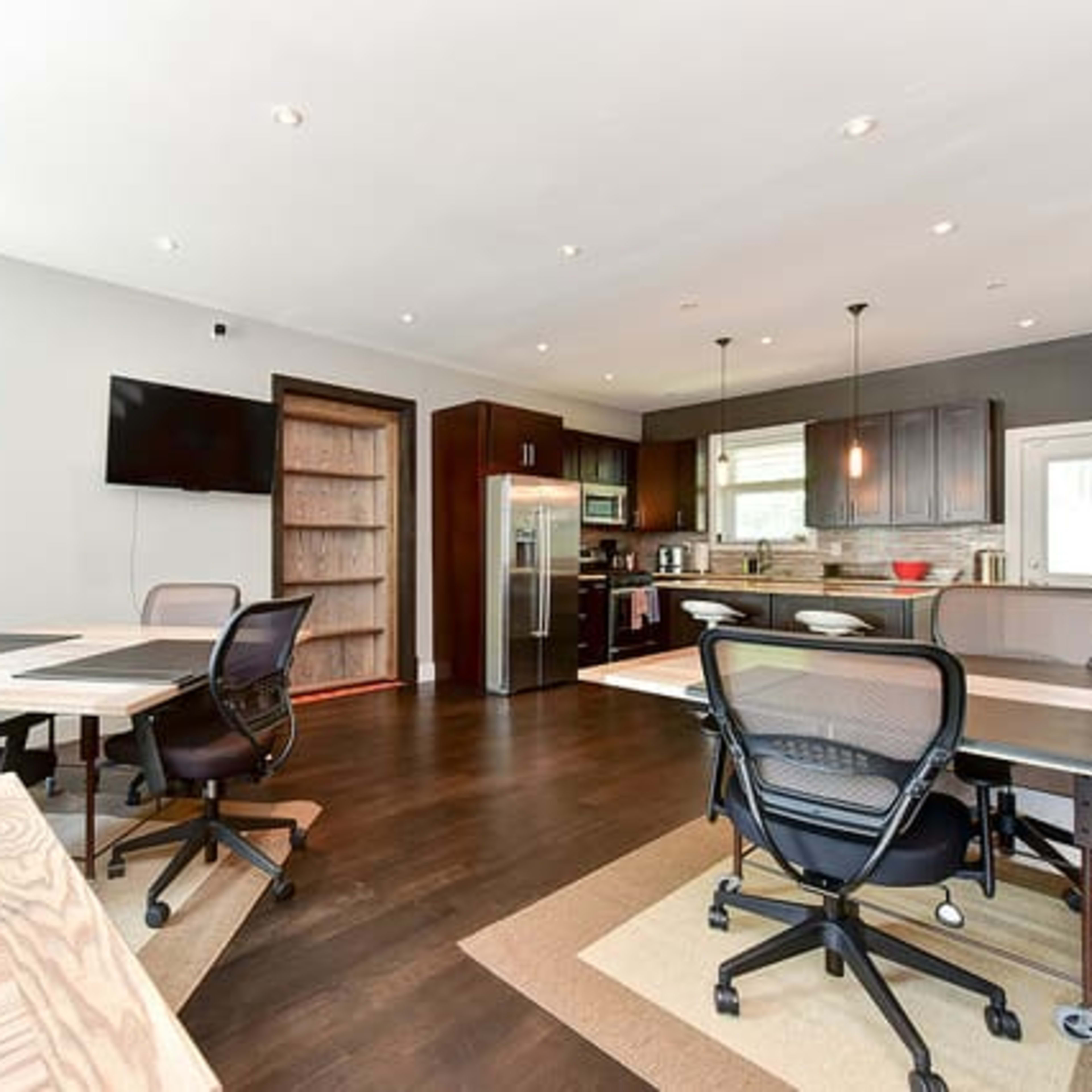 A modern office space with several work tables and chairs, a kitchenette area, and a large television mounted on the wall.