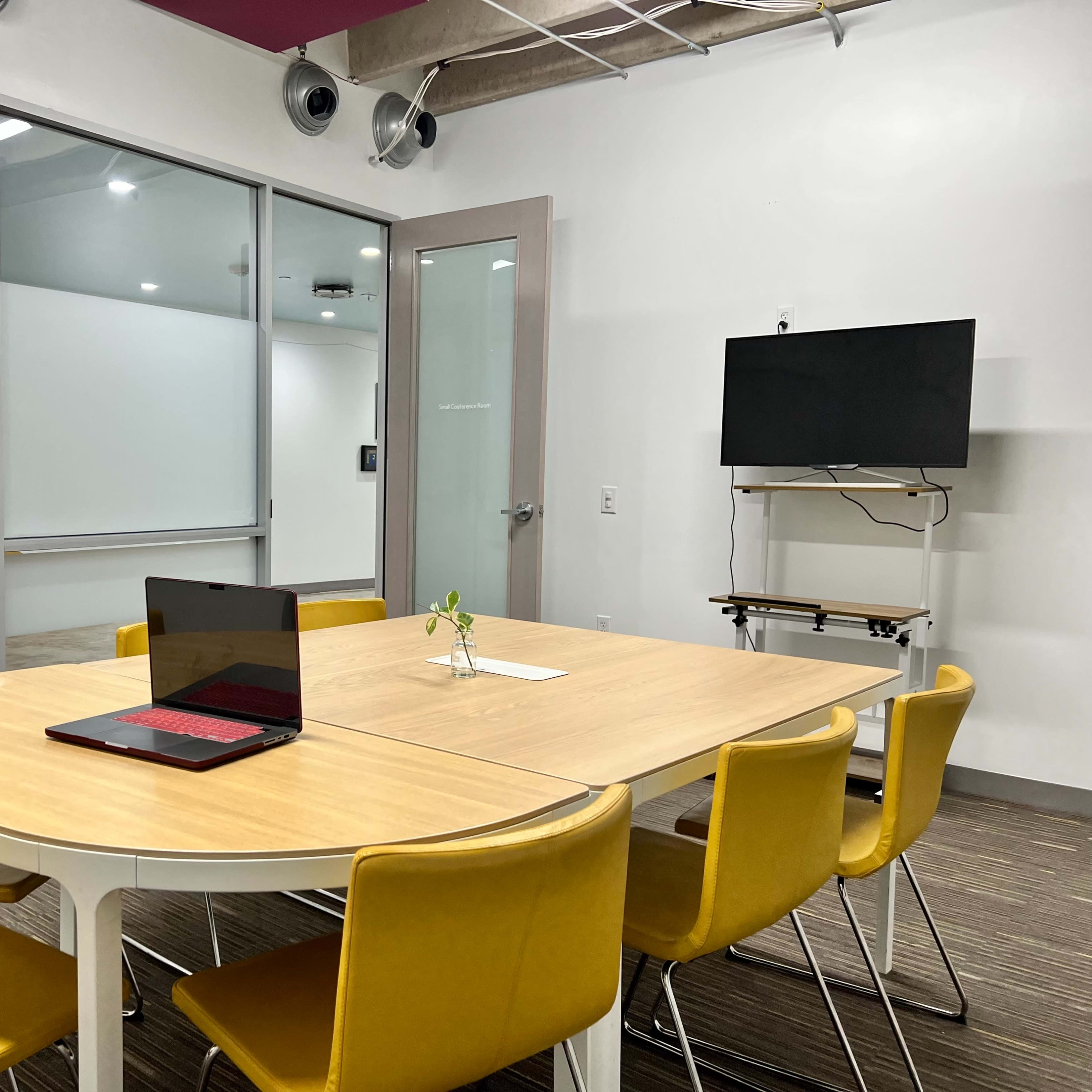 A conference room features a round table surrounded by yellow chairs, with a laptop open on the table and a television mounted on the wall.