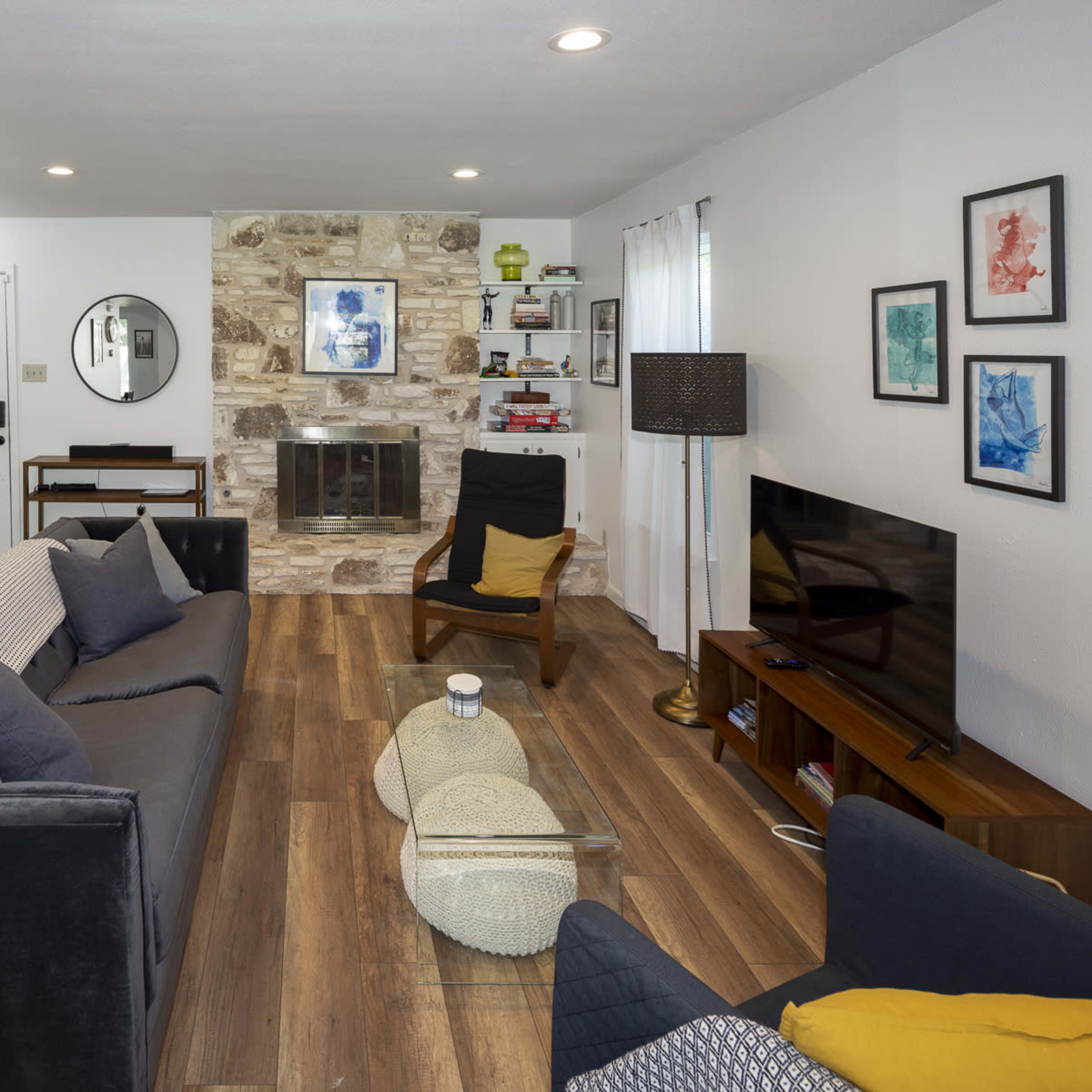 The living room features two sofas facing a coffee table, a stone fireplace, and a TV stand against a wall, with a front door and staircase visible in the background.