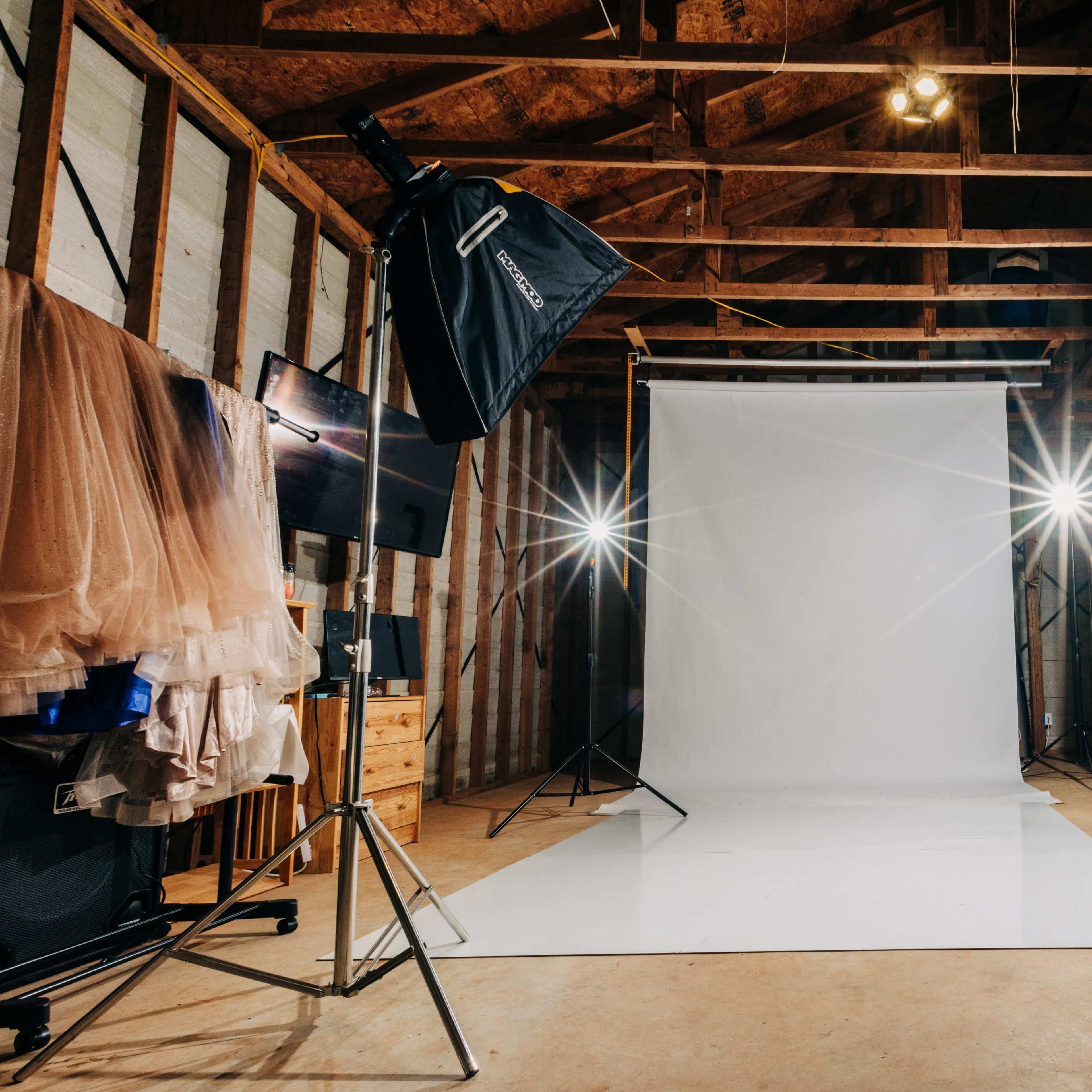 A photography studio setup with a white backdrop, lighting equipment, and various fabric drapes arranged in a rustic wooden barn interior.