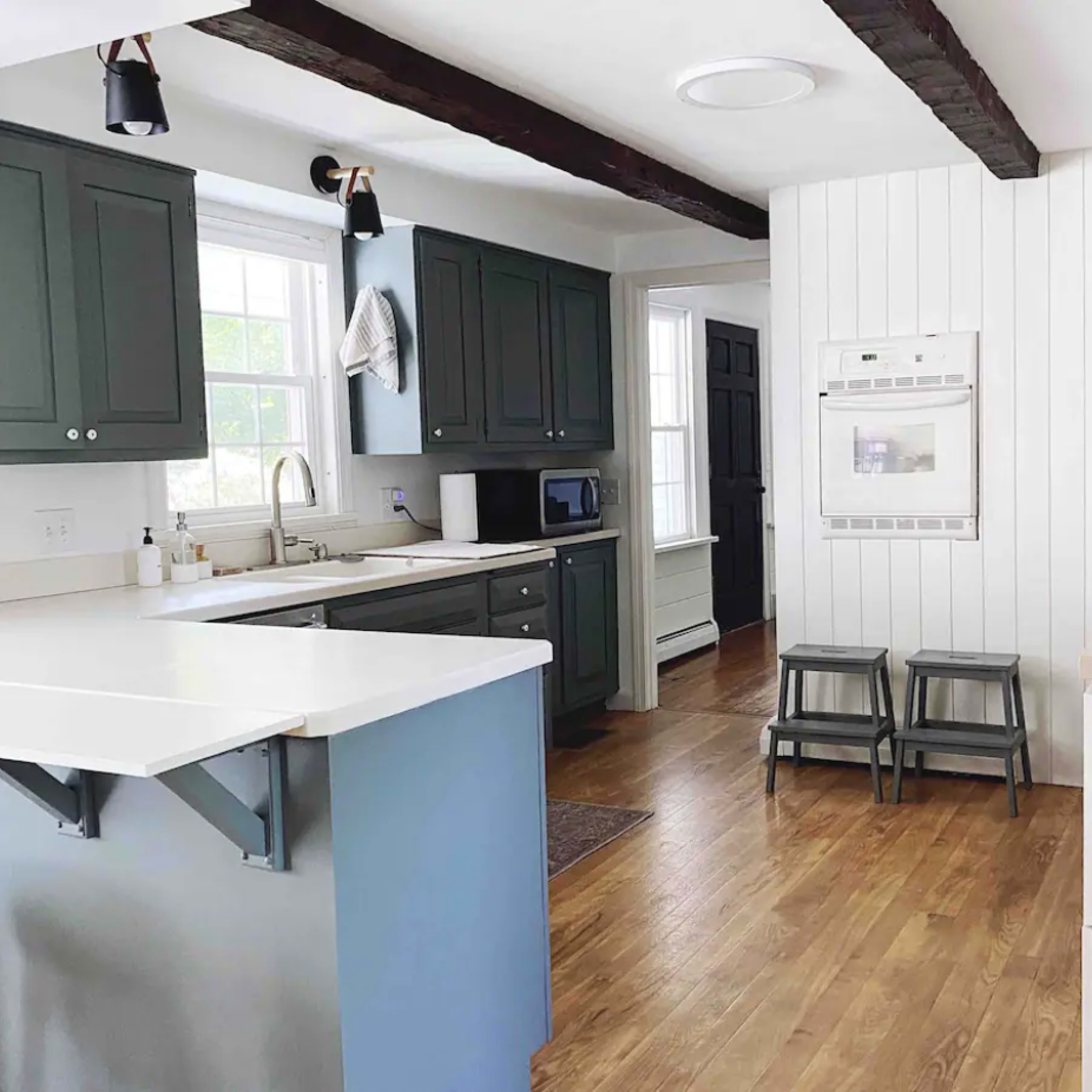 The kitchen features a combination of dark green and white cabinetry, stainless steel appliances, and wooden flooring, with a bar counter and two stools on one side.