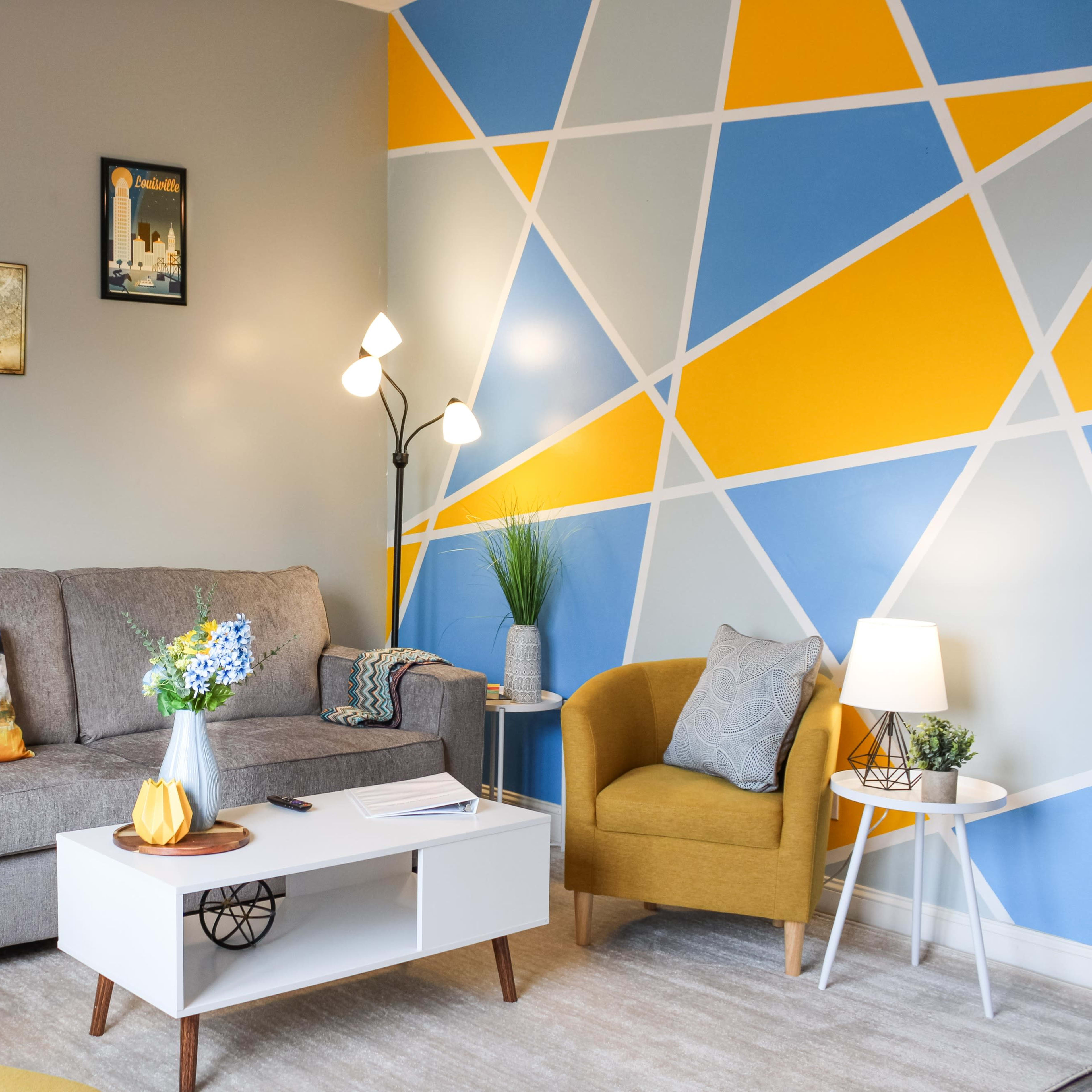 A modern living room with a gray sofa, two yellow chairs, a coffee table, and a colorful geometric wall design in blue and yellow.