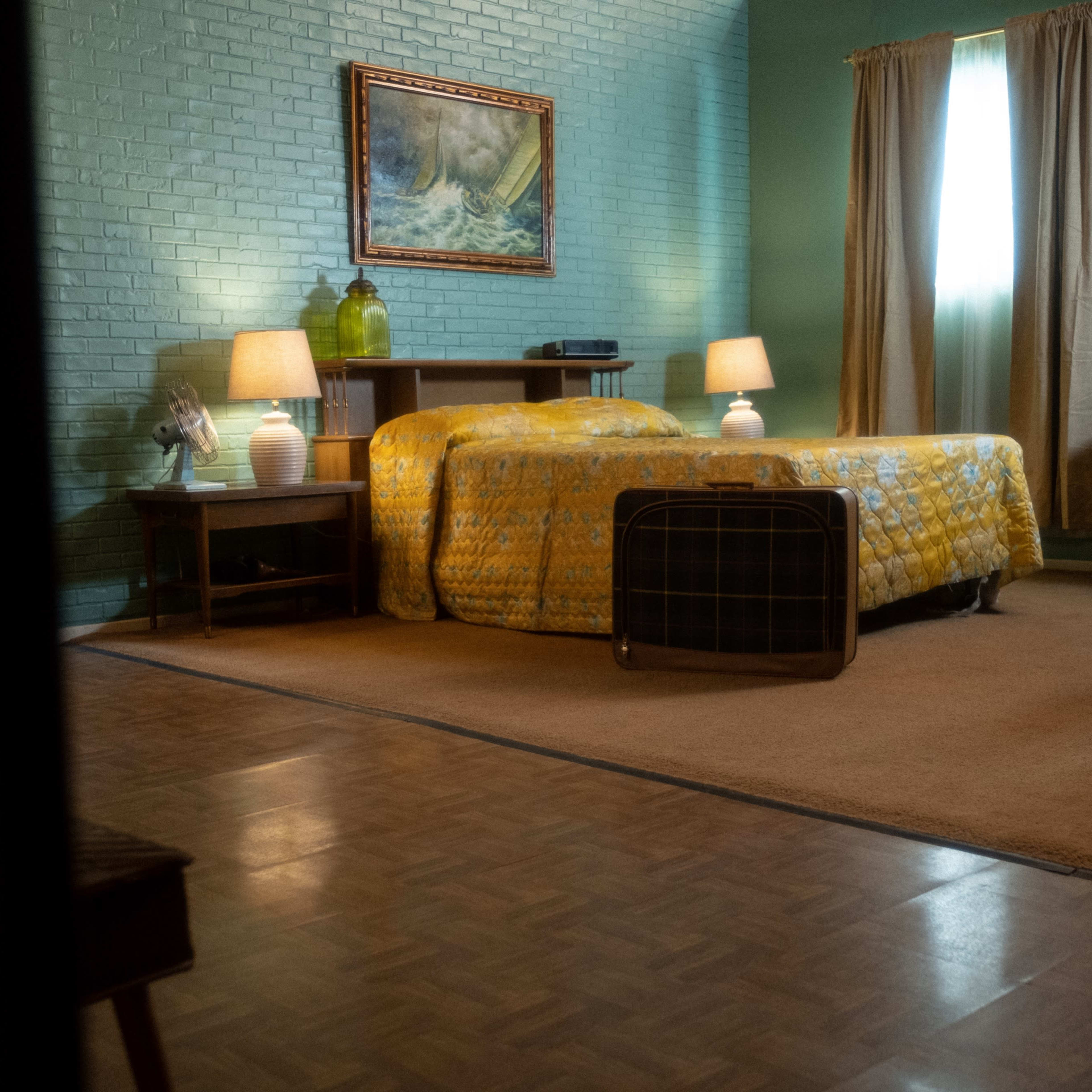 A neatly arranged bedroom features a bed with a yellow patterned blanket, two lamps on bedside tables, and a suitcase on the floor near a window with curtains.