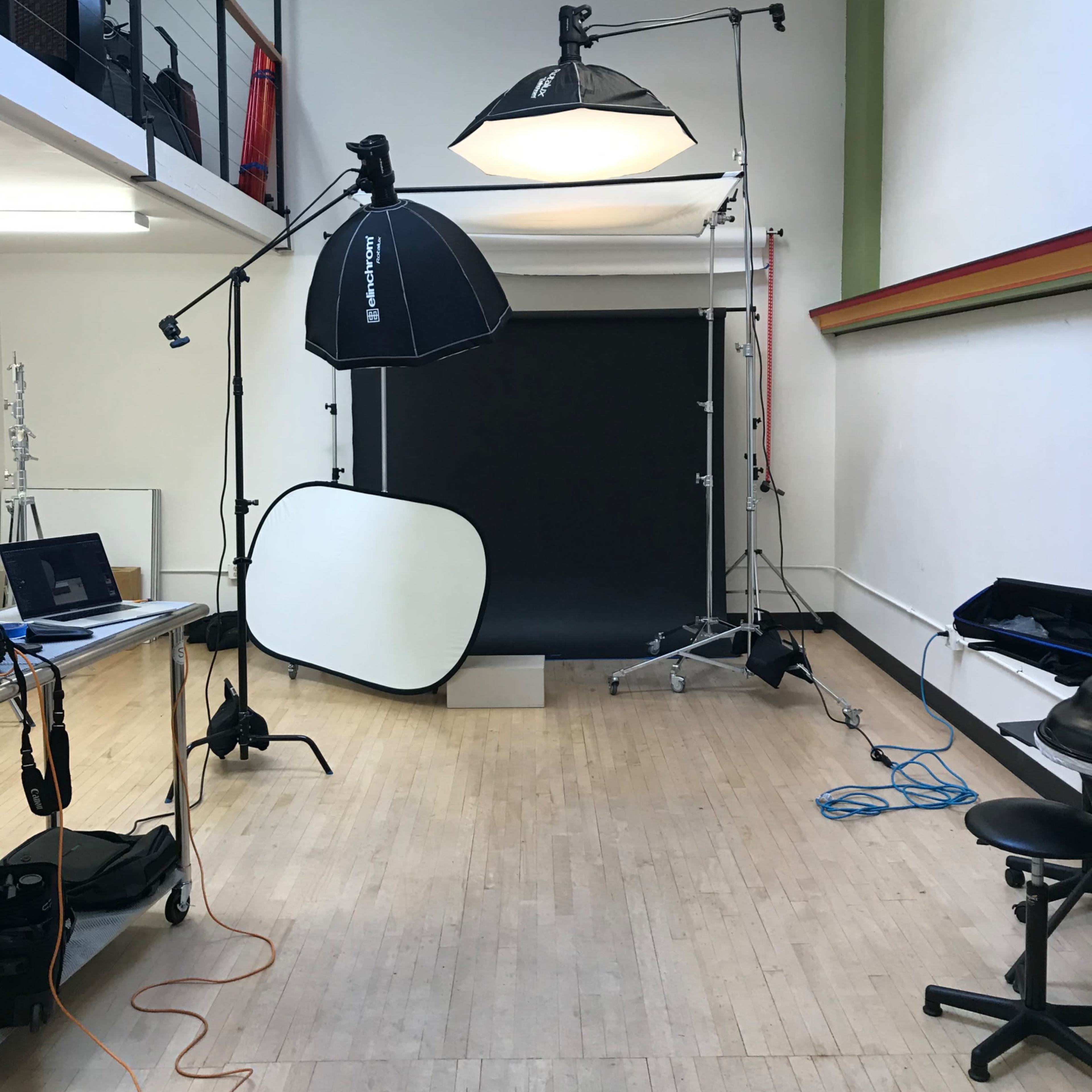 A photography studio is set up with lighting equipment, a dark backdrop, and various props arranged on the floor.
