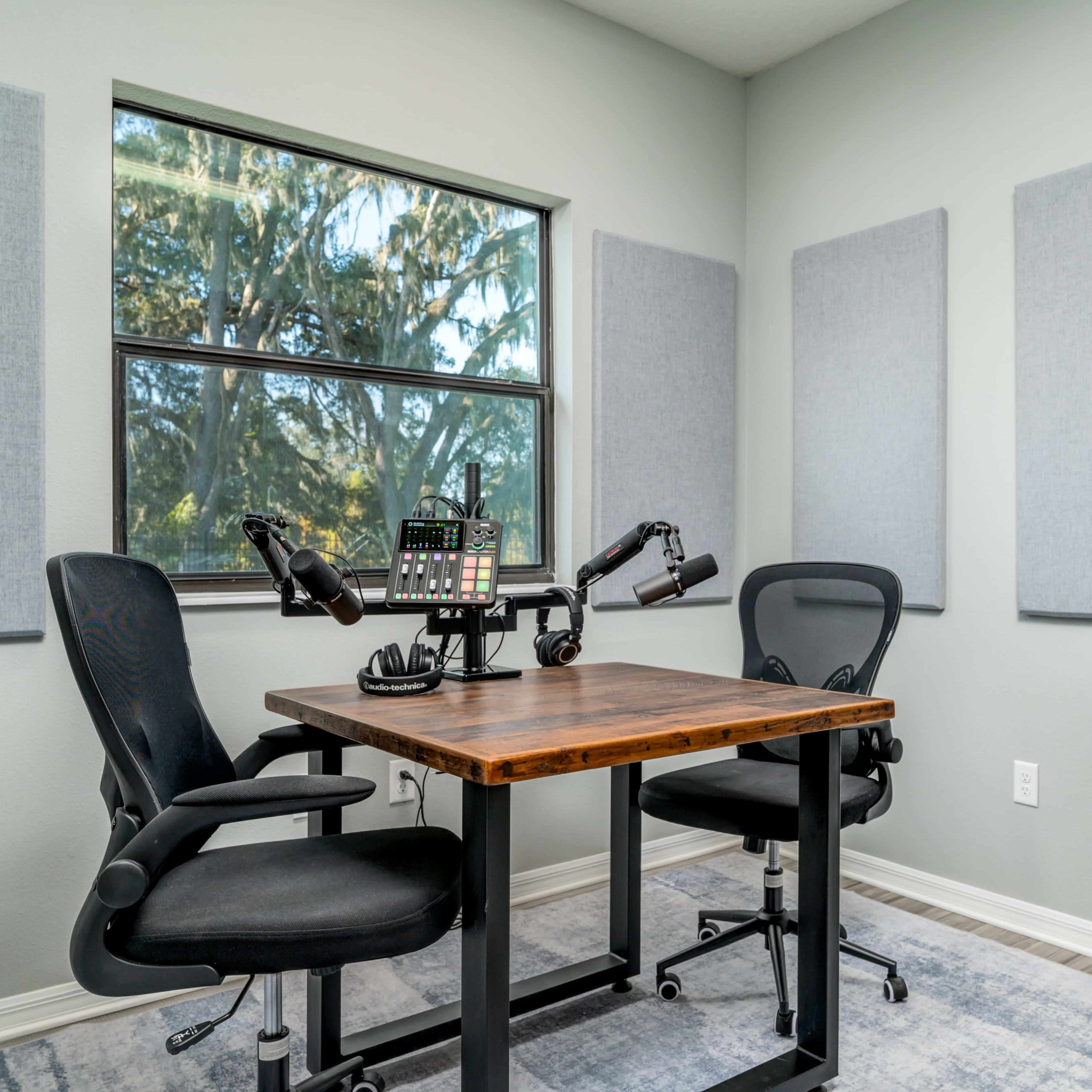 A small podcasting room with a wooden table and two black chairs, featuring soundproofing panels on the walls and a microphone setup on the table.