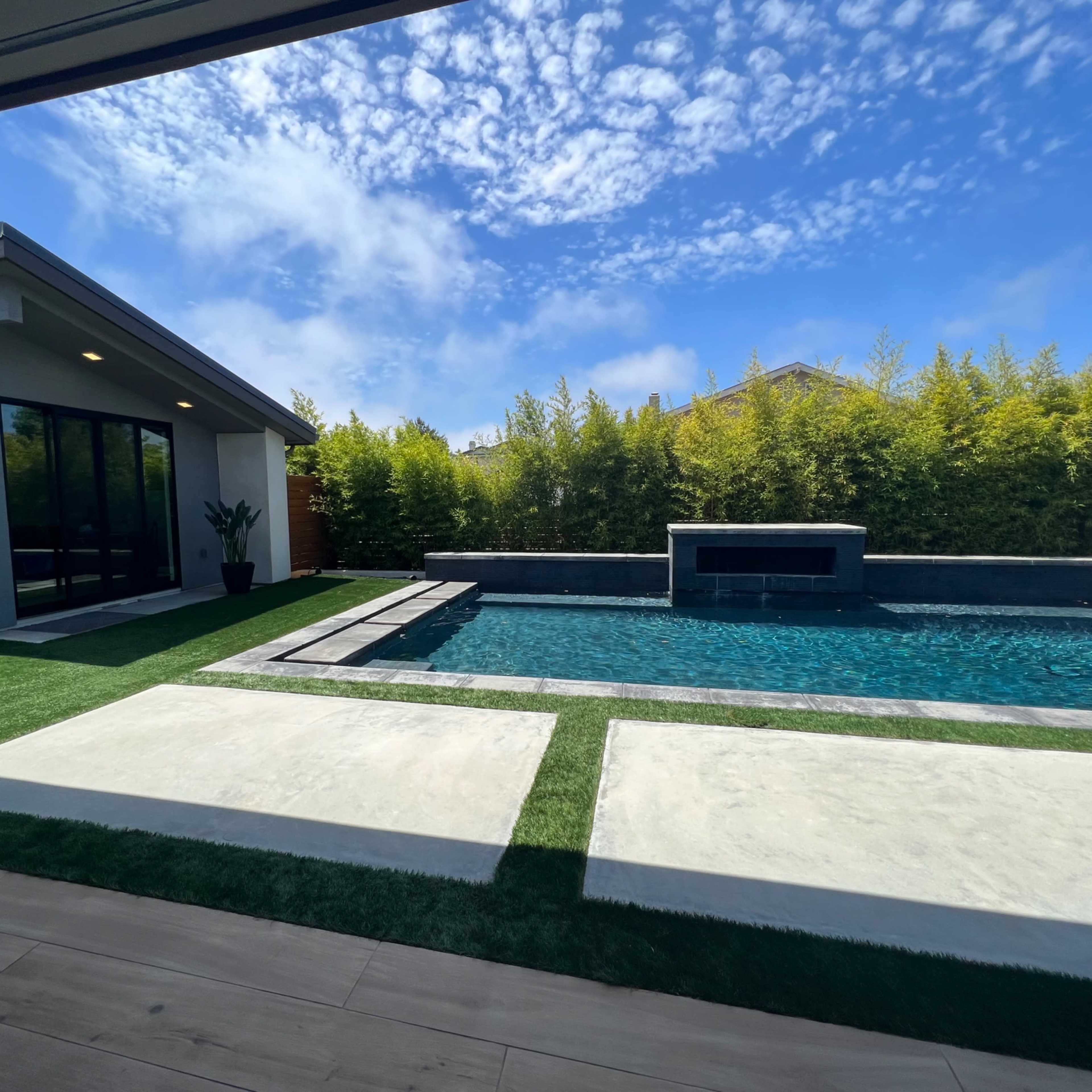 A modern backyard with a pool, concrete patio, and lush greenery surrounding a contemporary house under a blue sky.