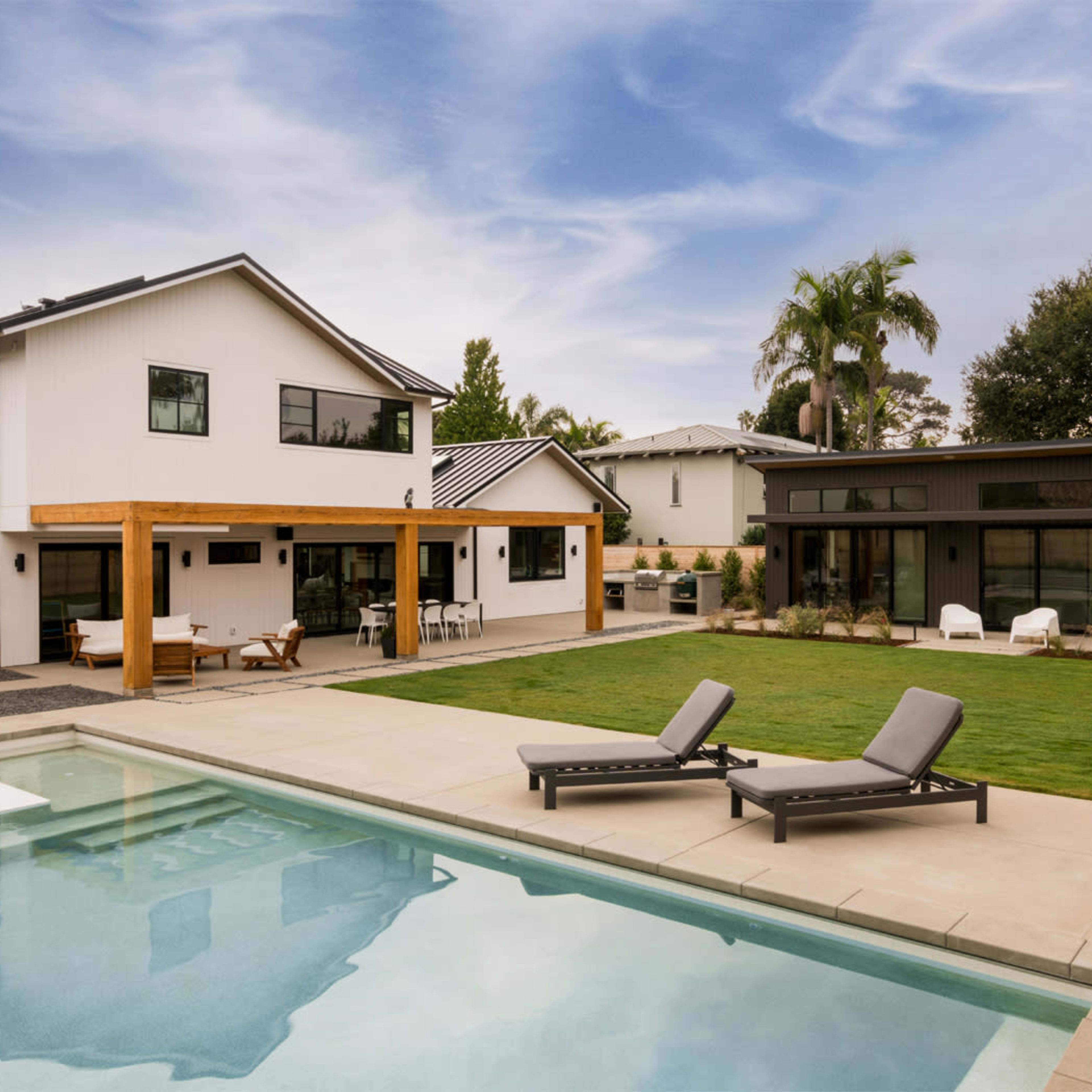 A modern backyard features a swimming pool, lounge chairs, and a stylish home with a wooden pergola and landscaped grass areas.