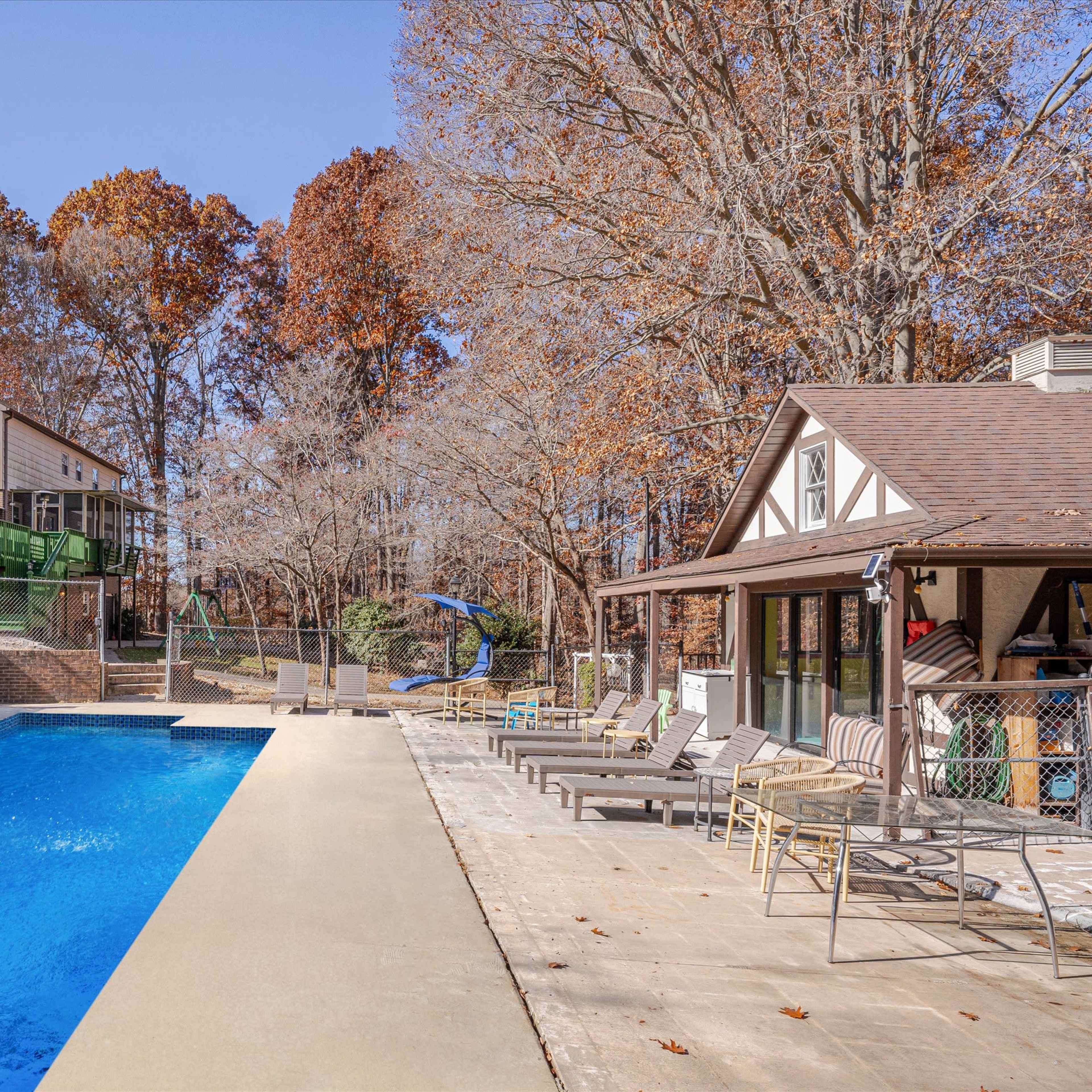 A swimming pool surrounded by concrete and chairs, with two houses in the background and trees with autumn foliage.