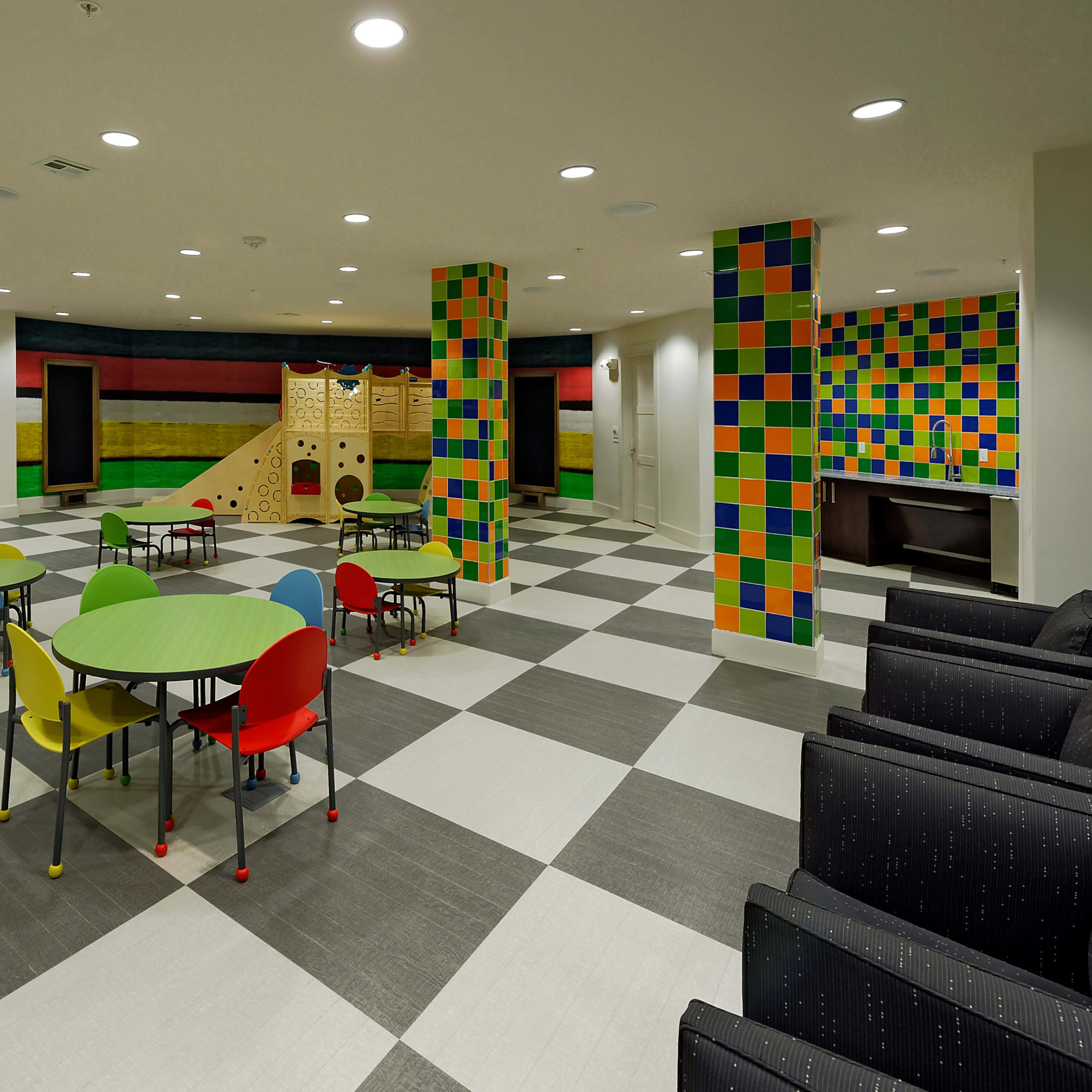 A brightly colored playroom with multicolored wall panels, green and red tables, and a climbing structure.