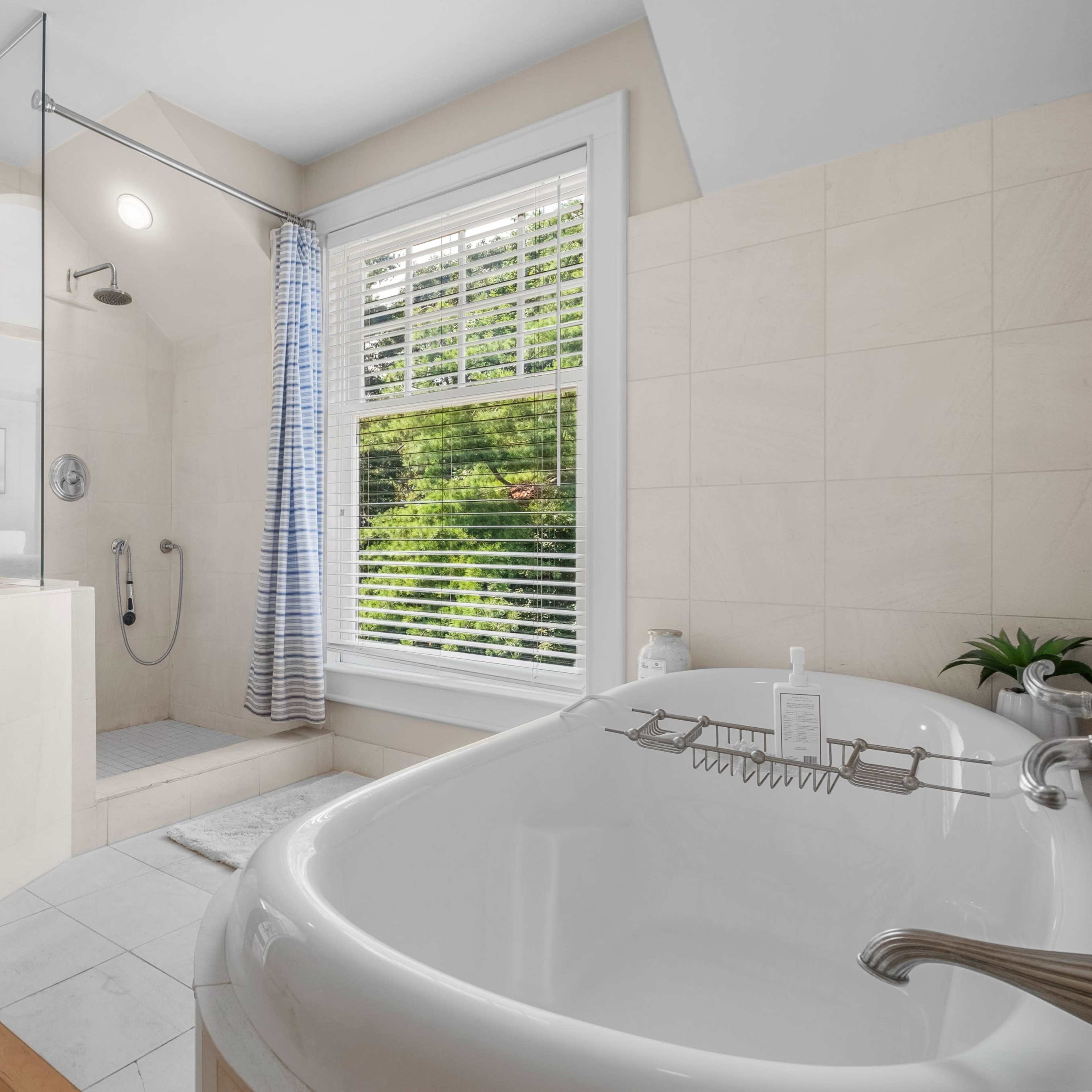 A modern bathroom featuring a glass shower, a freestanding bathtub, and large windows providing natural light and a view of greenery outside.